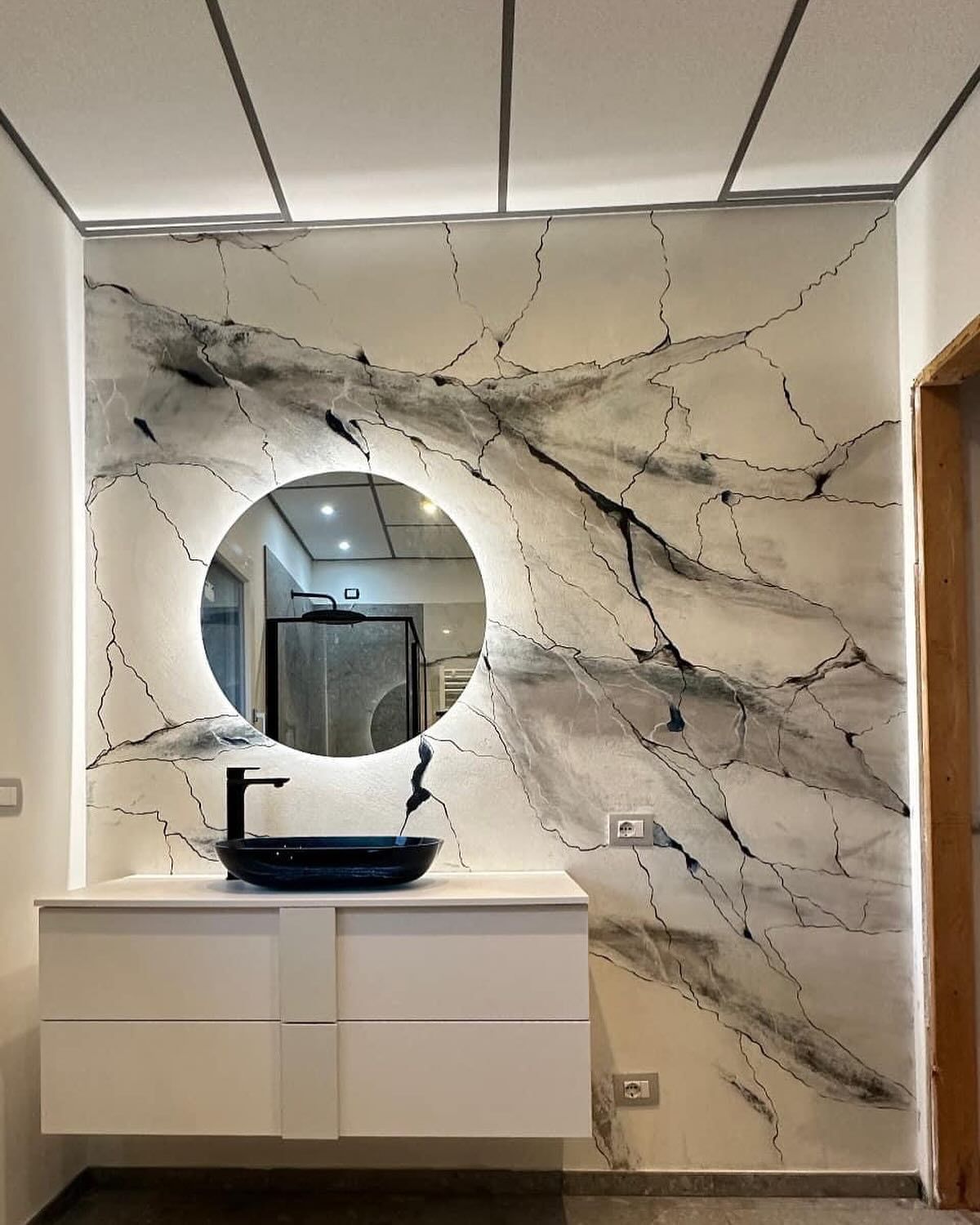 Mobile bagno moderno con lavabo a ciotola nero e specchio rotondo retroilluminato, su una parete bianca con venature in marmo scuro.