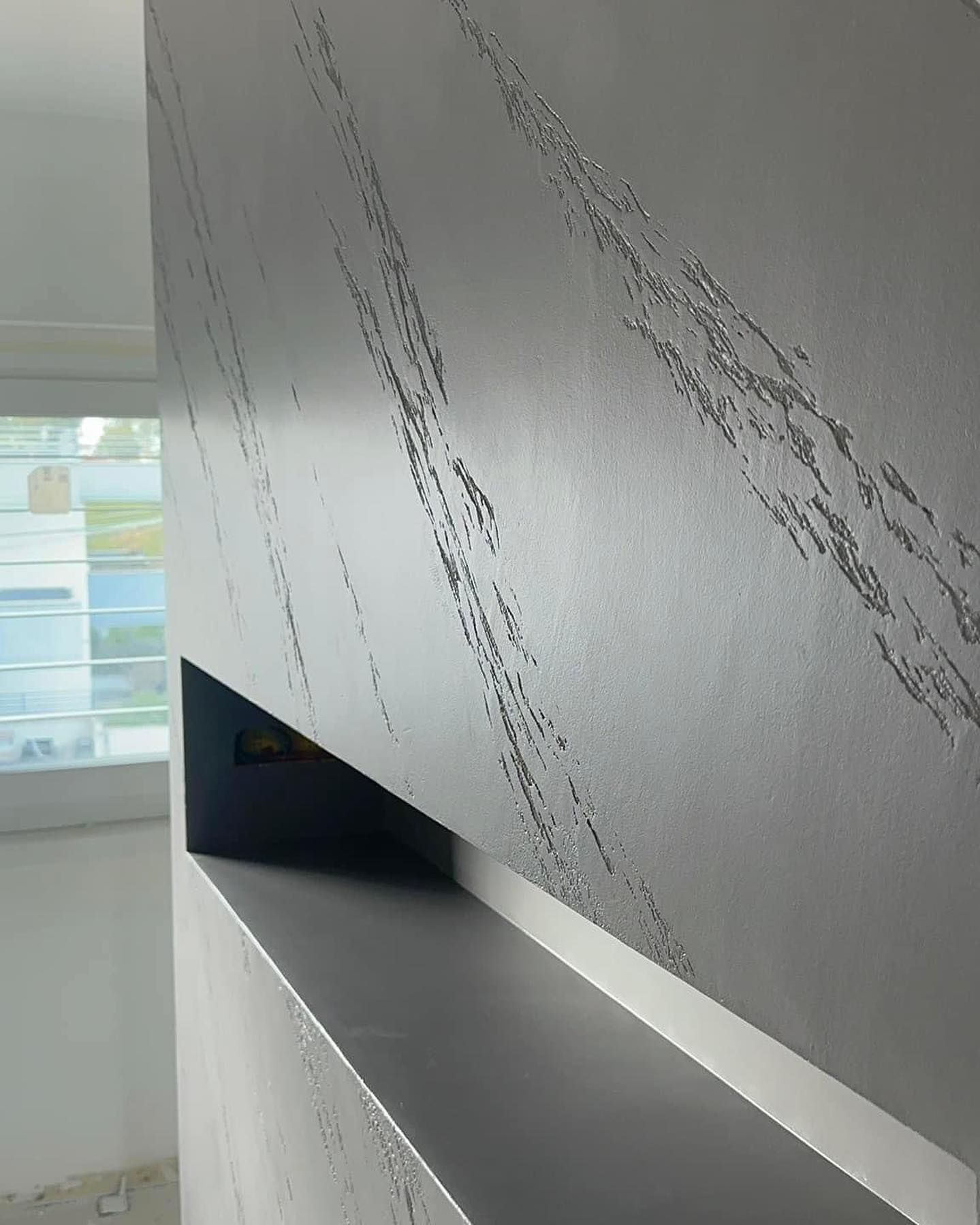 Primo piano di una parete grigia e ruvida con striature diagonali scintillanti e una mensola orizzontale nera integrata.