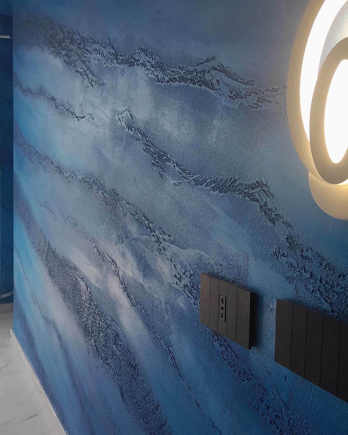 Una parete blu con una finitura di pittura ondulata e ruvida e due placche rettangolari nere per interruttori elettrici sulla destra.