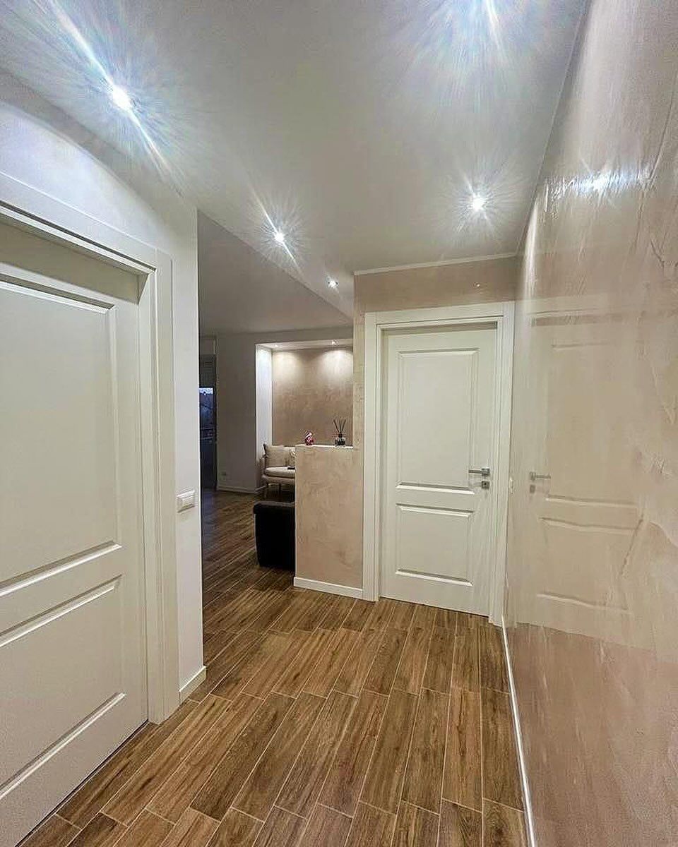 Un corridoio con pavimento effetto legno, porte interne bianche e pareti beige con finitura ruvida, illuminate da faretti a incasso sul soffitto.