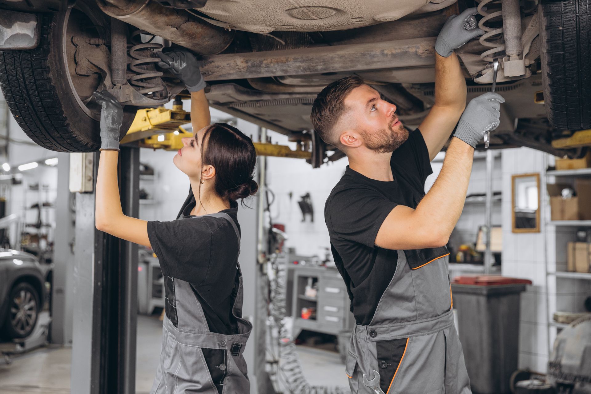 Two mechanics, wearing grey overalls, working under a car in a garage.