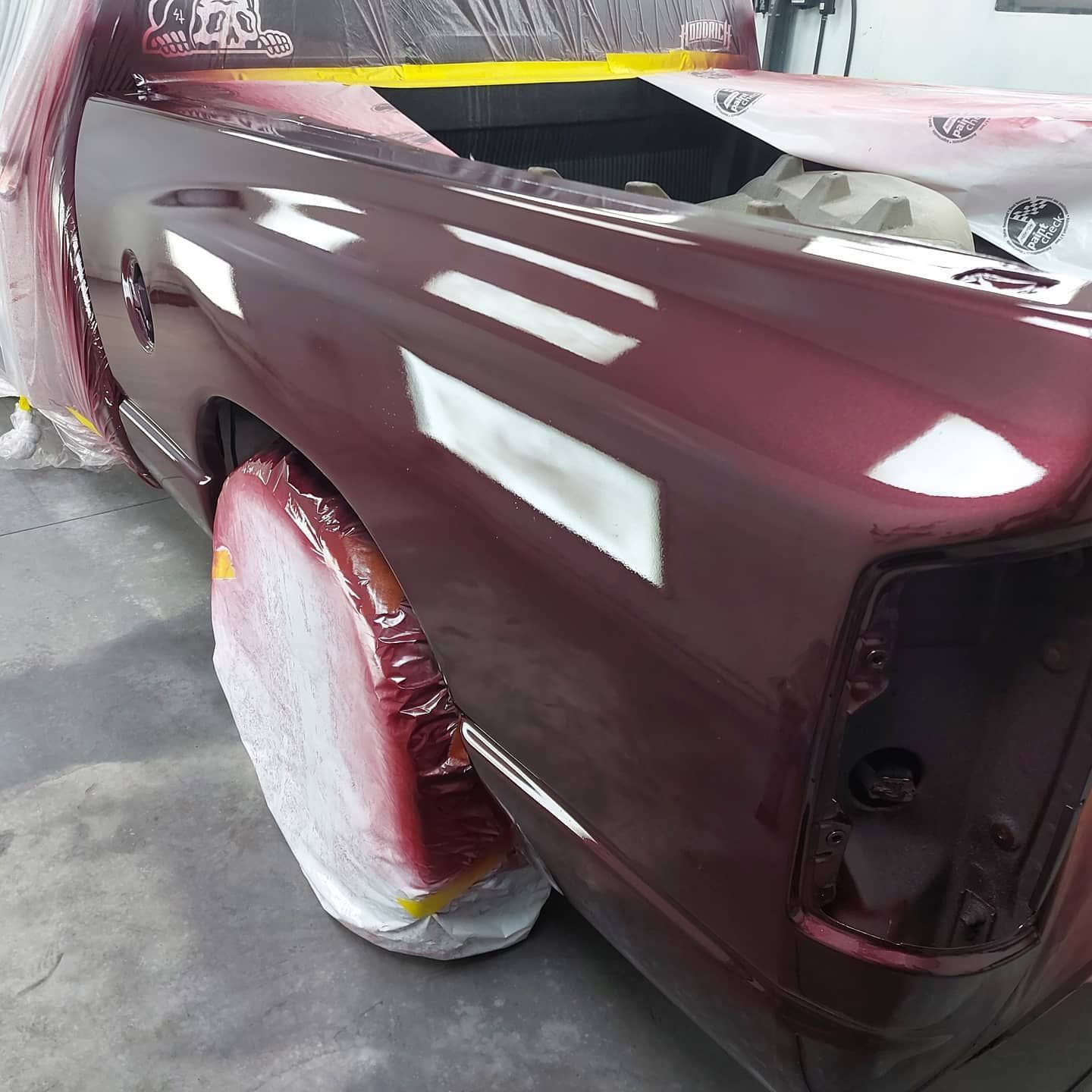 Maroon car body in a paint shop, with masked wheel and panel edges, under bright fluorescent lights