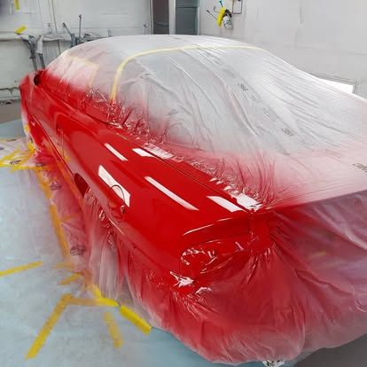 Car partially covered in plastic, freshly painted bright red in a paint booth.