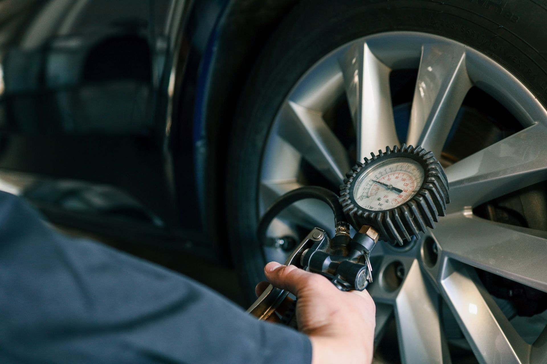 A person checks tire pressure with a gauge near a car's wheel in a shop.