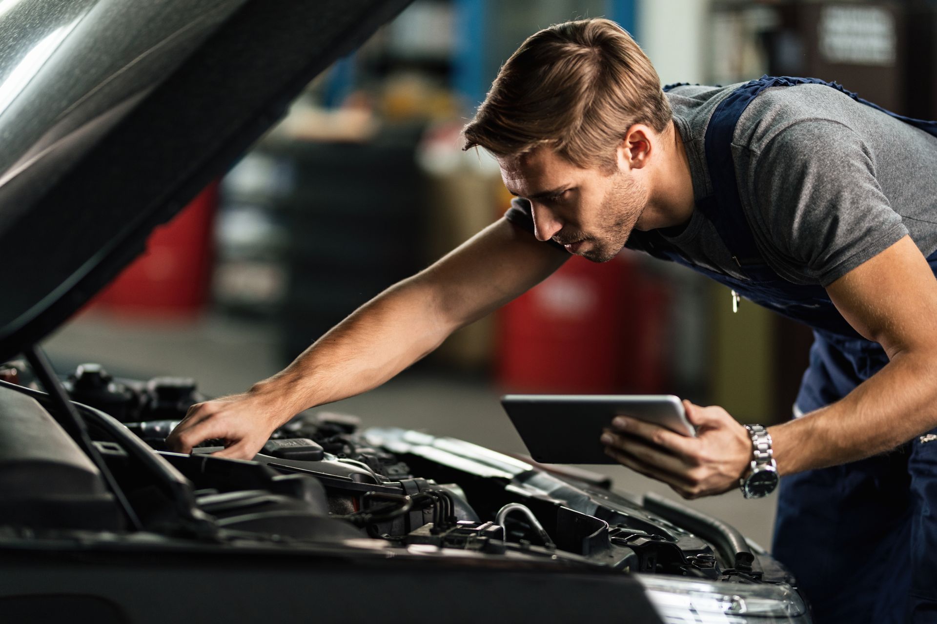 Mechanic examining car engine while holding a tablet in a garage.