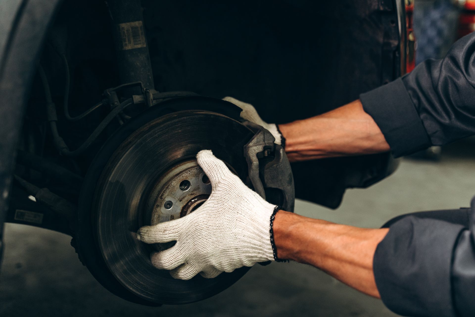 Mechanic in gloves working on a car brake, holding the disc with both hands in a garage setting.