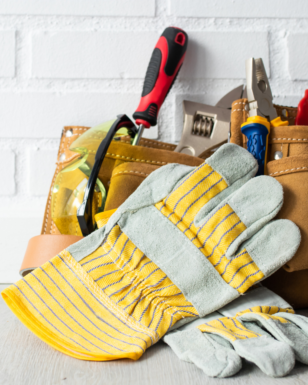 Des gants de travail posés devant une ceinture à outils en cuir beige contenant un tournevis, une pince et une clé réglable.