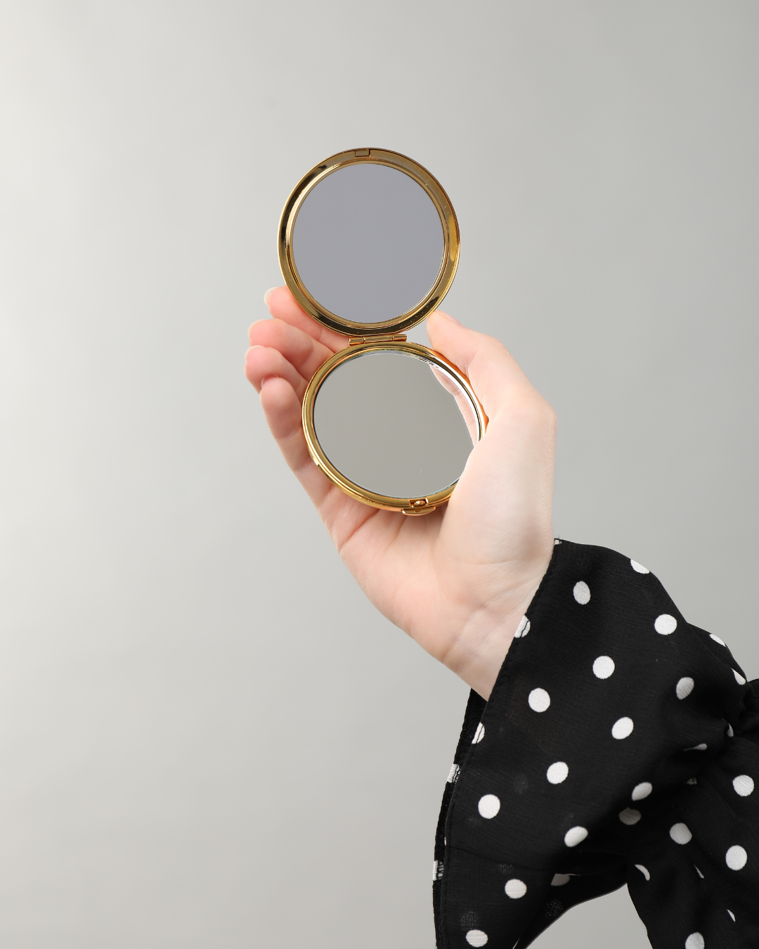Sur un fond clair, une main vêtue d'une manche à pois noirs et blancs tient ouvert un miroir de poche rond à bordure dorée.