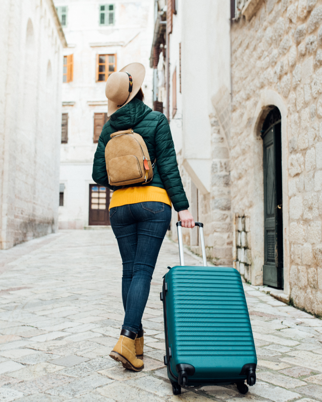 Une personne portant un chapeau, une veste verte et un jean marche dans une rue pavée étroite, tirant une valise turquoise