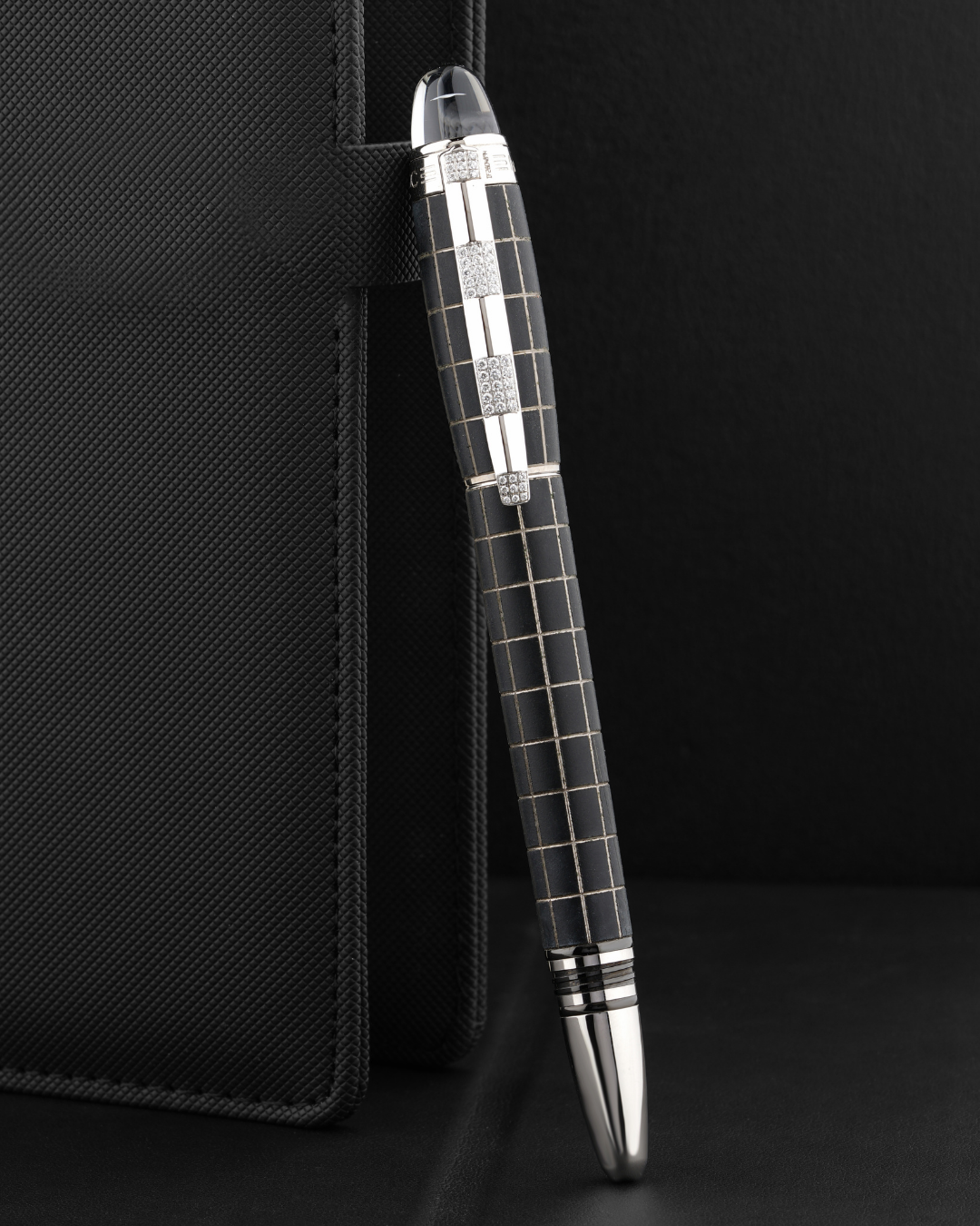 Un stylo noir à motif quadrillé, avec un clip et une pointe argentés, repose contre le côté d'un cahier noir fermé.