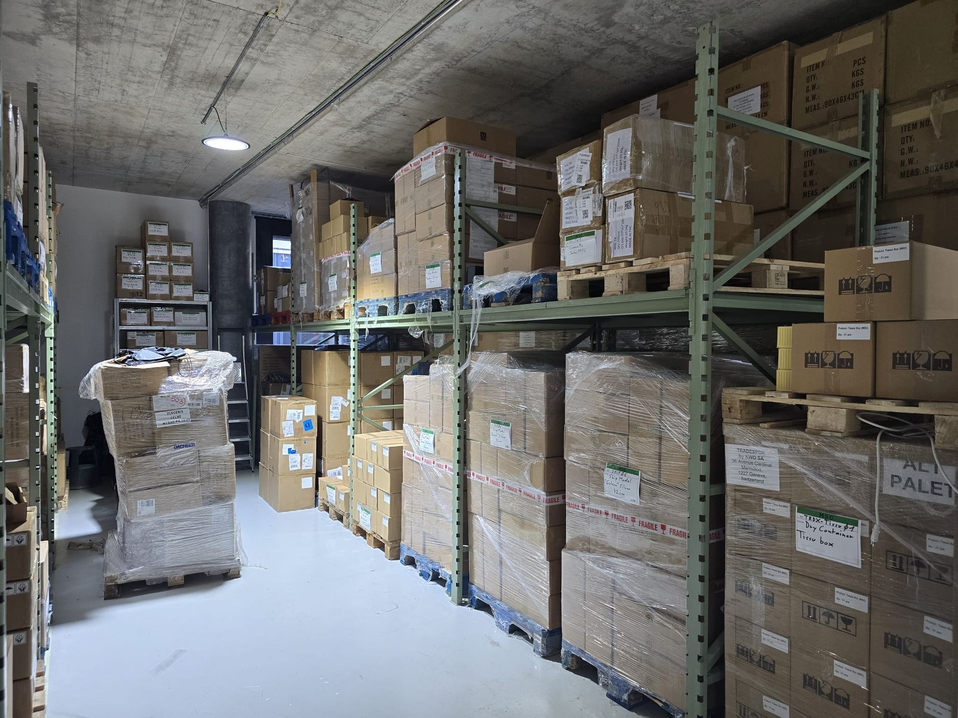 L'intérieur d'un entrepôt bien éclairé présente de hautes étagères métalliques remplies de nombreux cartons sur des palettes.