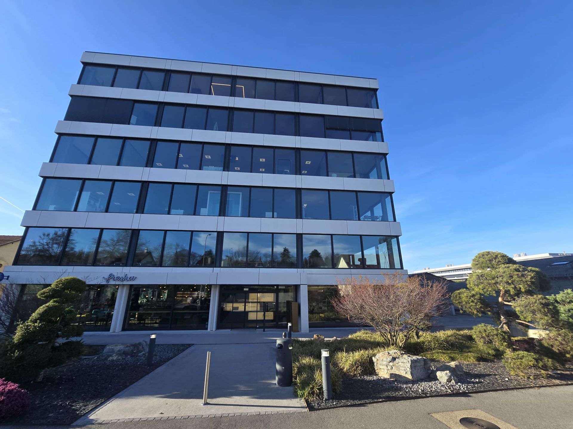Un immeuble de bureaux de cinq étages avec une façade en verre, des moulures horizontales blanches et une entrée paysagée