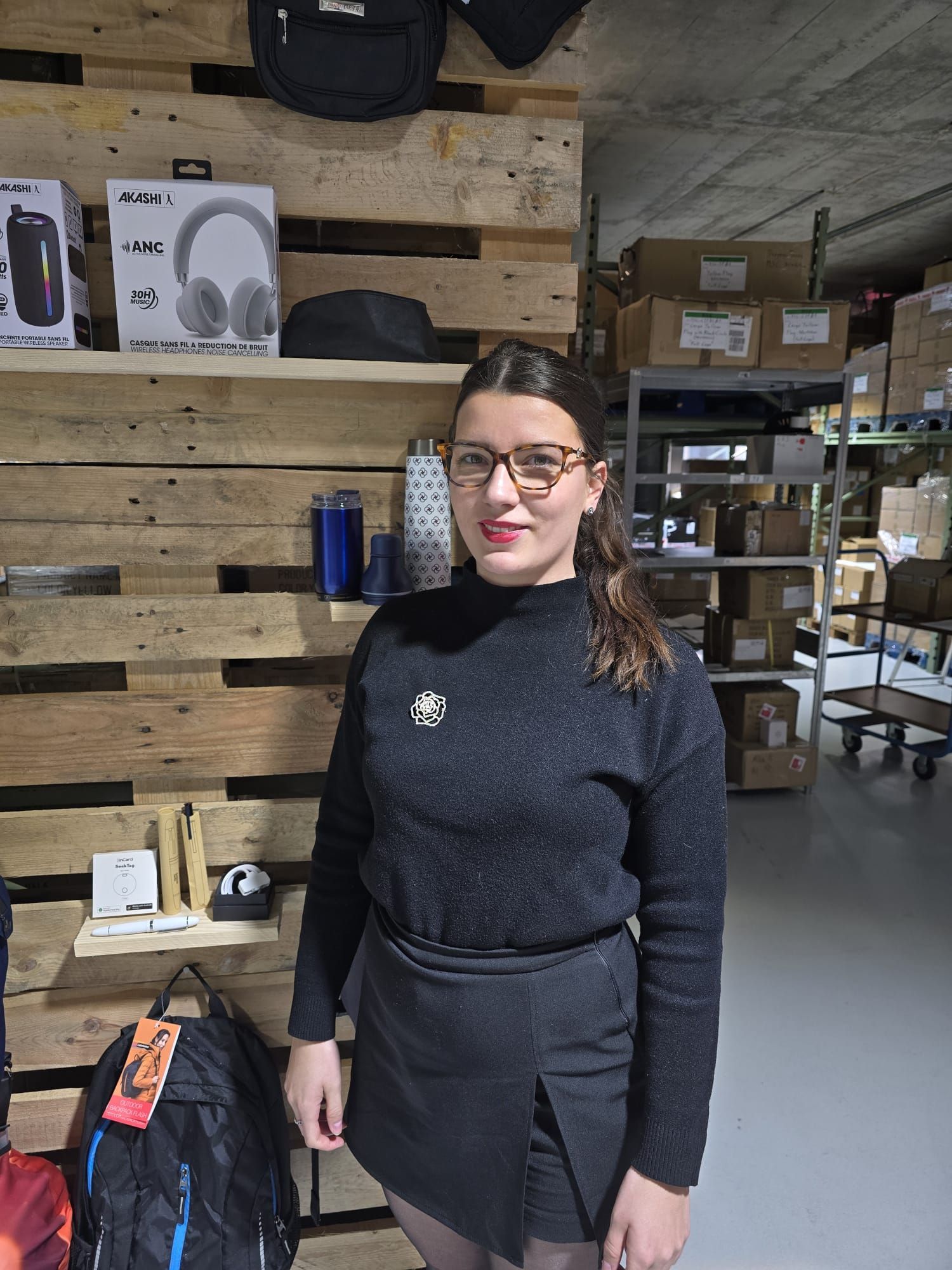 Une personne portant des lunettes et un pull noir se tient dans un entrepôt devant un présentoir en palettes de bois