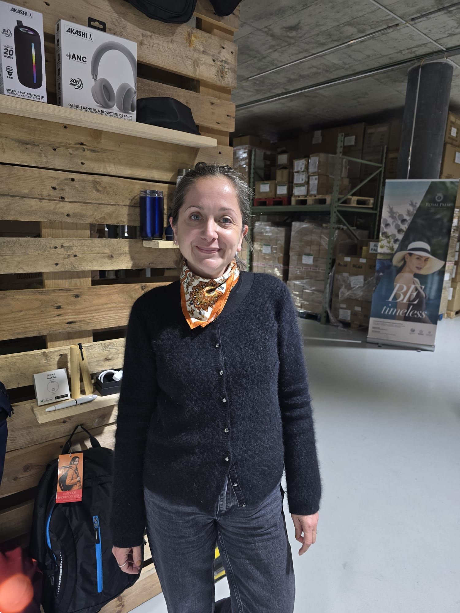 Une personne vêtue d'un pull noir et d'une écharpe à motifs sourit, debout dans un entrepôt avec des étagères en bois