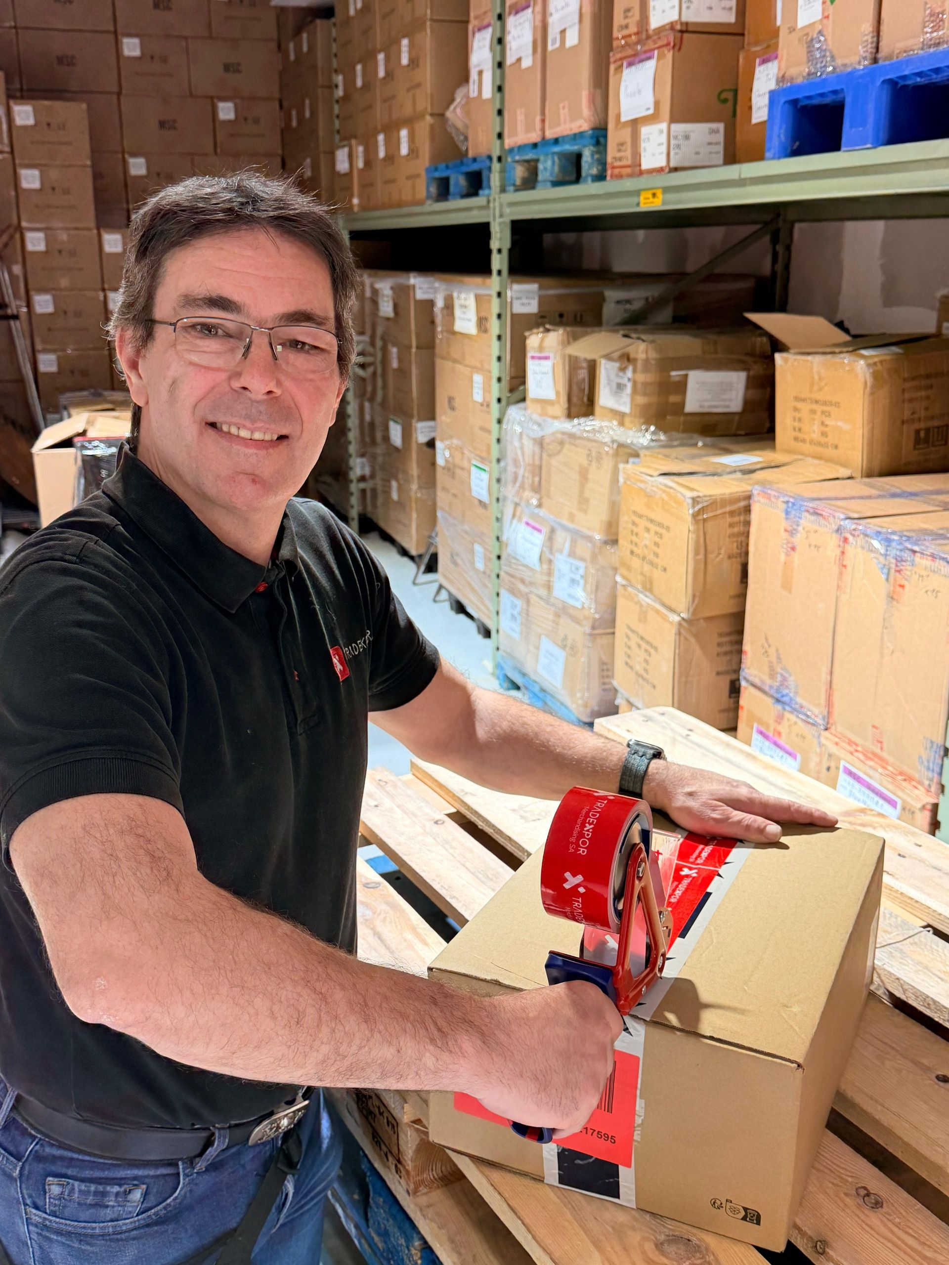 Une personne vêtue d'un polo noir sourit en utilisant un dérouleur de ruban adhésif rouge pour sceller une boîte en carton