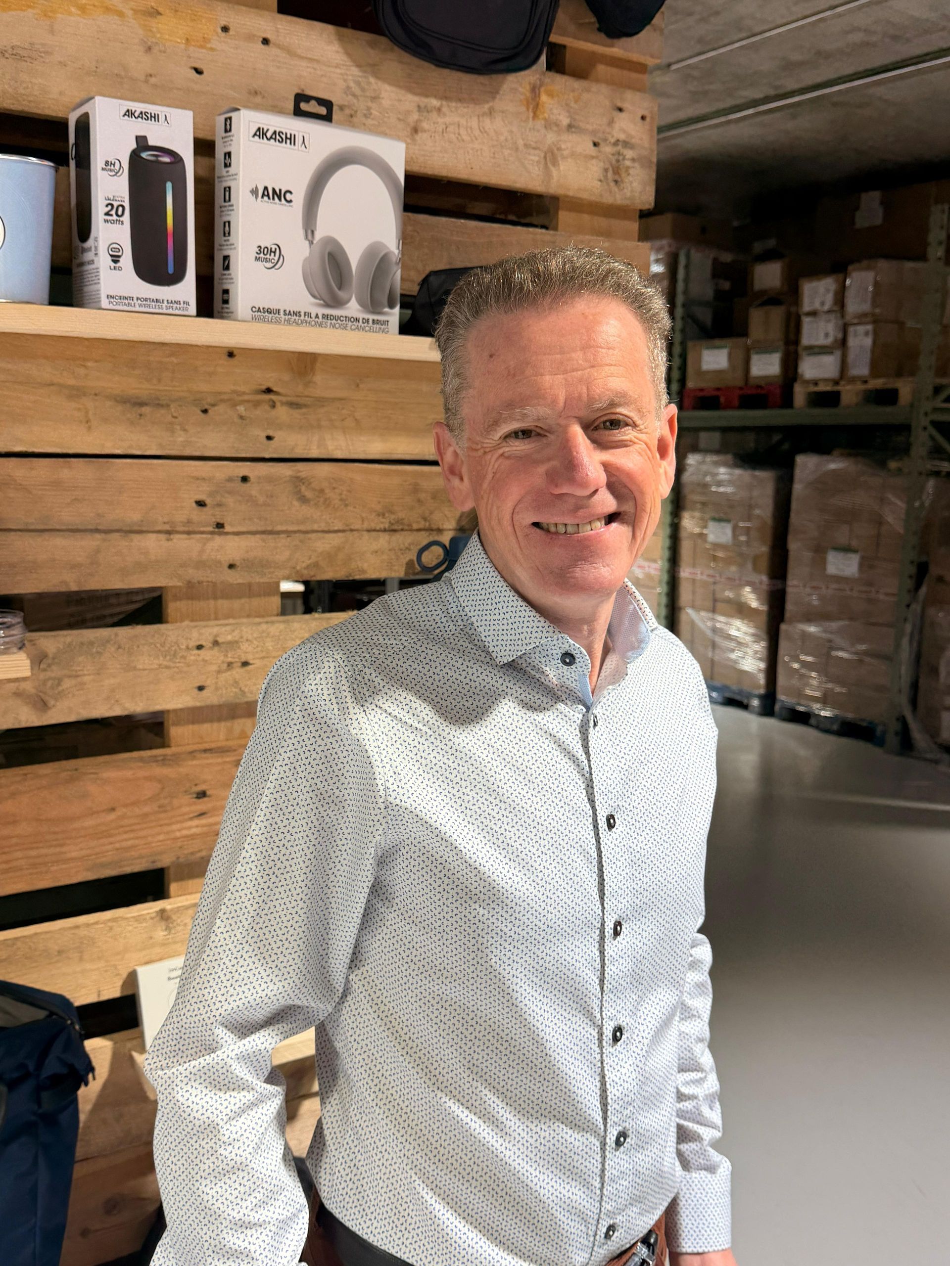 Une personne vêtue d'une chemise à motifs sourit devant des étagères remplies de produits électroniques.