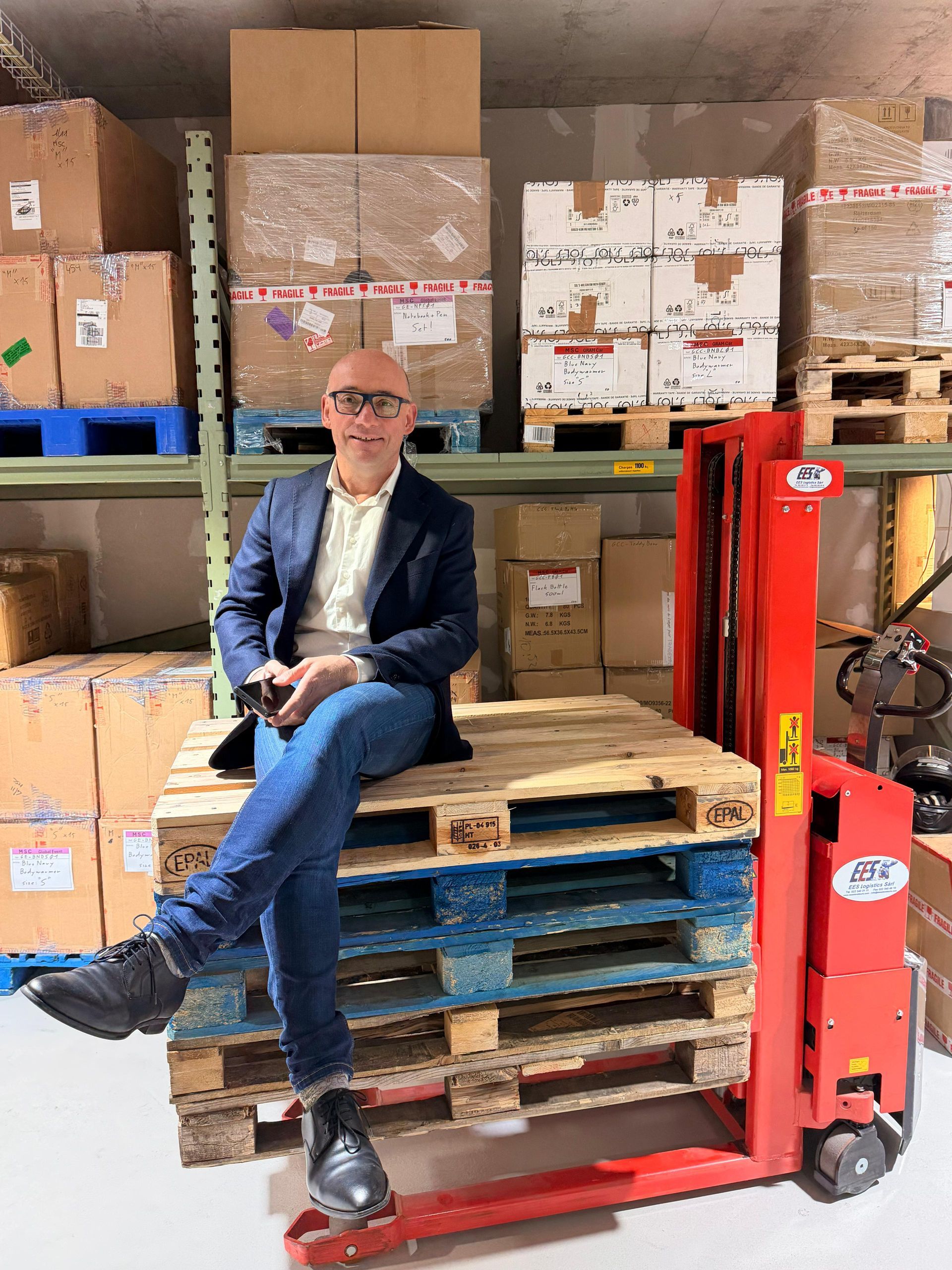 Dans un entrepôt, une personne vêtue d'un blazer et d'un jean est assise sur des palettes de transport en bois empilées, à côté d'un chariot