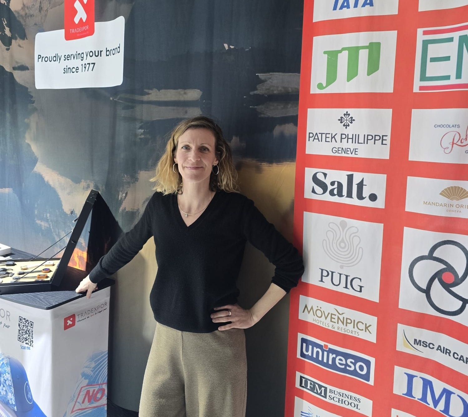 Une personne se tient devant une grande banderole événementielle affichant des logos d'entreprises, avec un stand promotionnel à proximité.