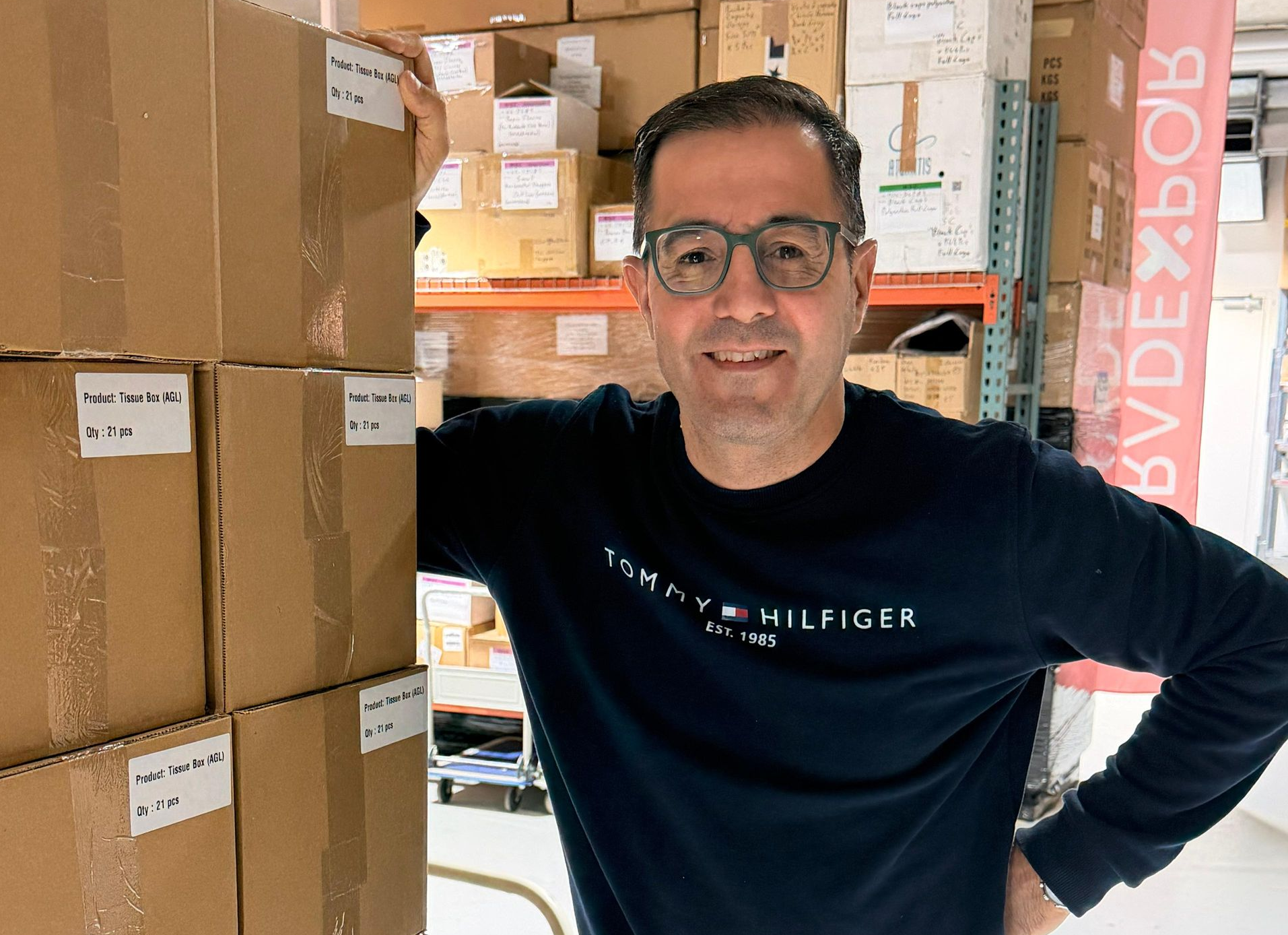 Un homme portant des lunettes et un sweat-shirt foncé se tient debout, souriant, dans un entrepôt