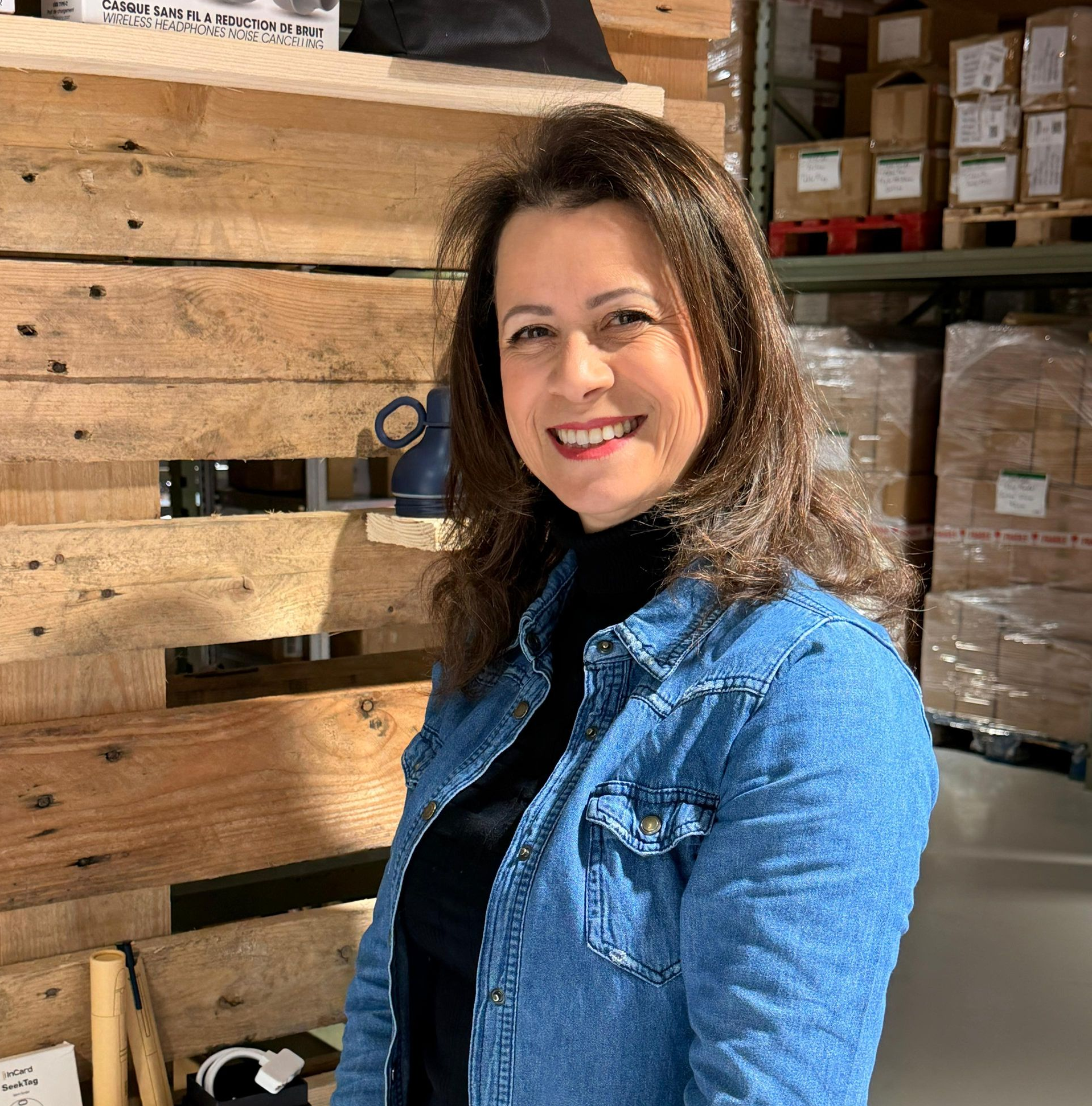 Une personne souriante, vêtue d'une chemise en jean par-dessus un col roulé noir, se tient debout dans un entrepôt