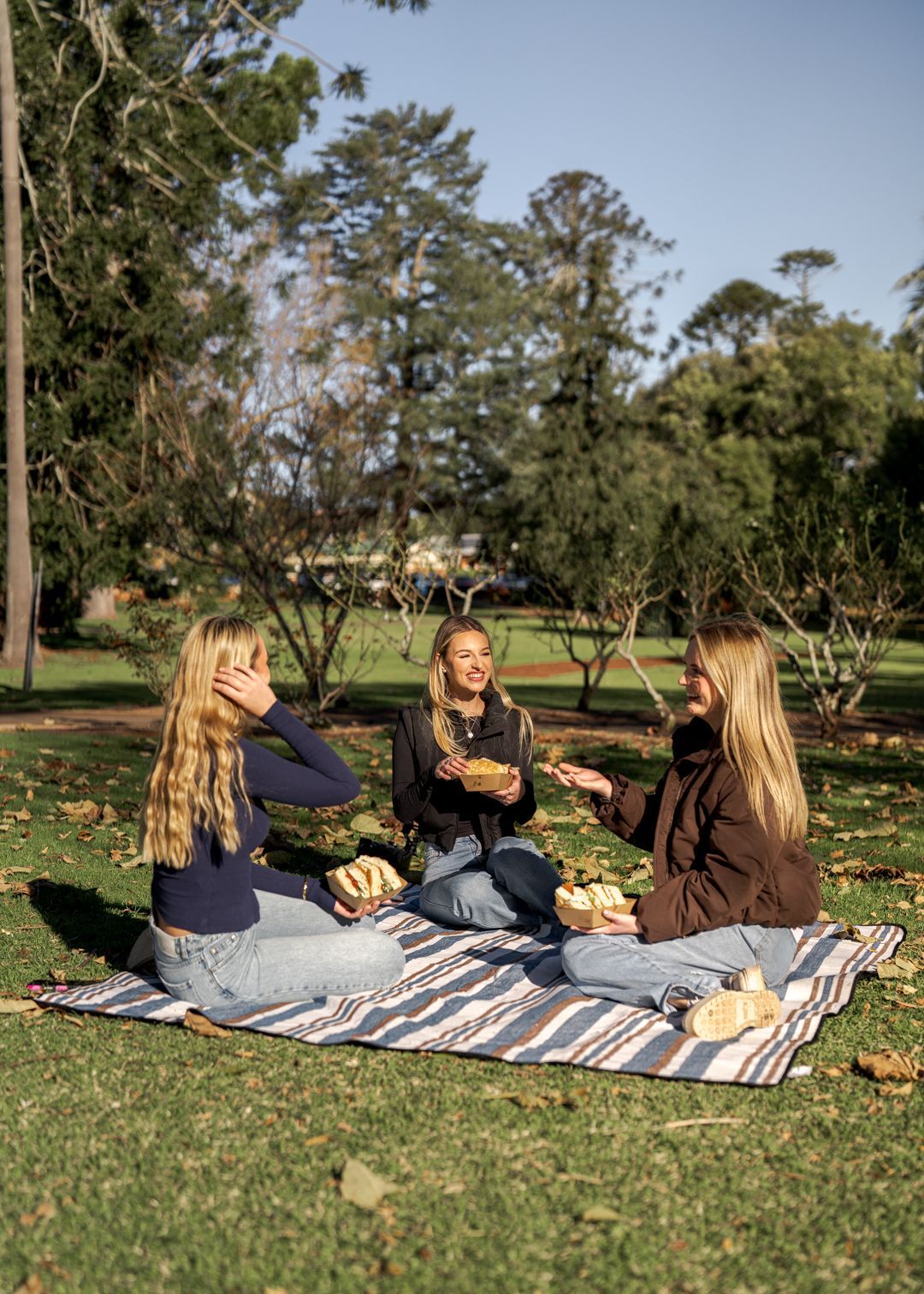 Three girls sitting on a picnic rug int he park with sandwiches 