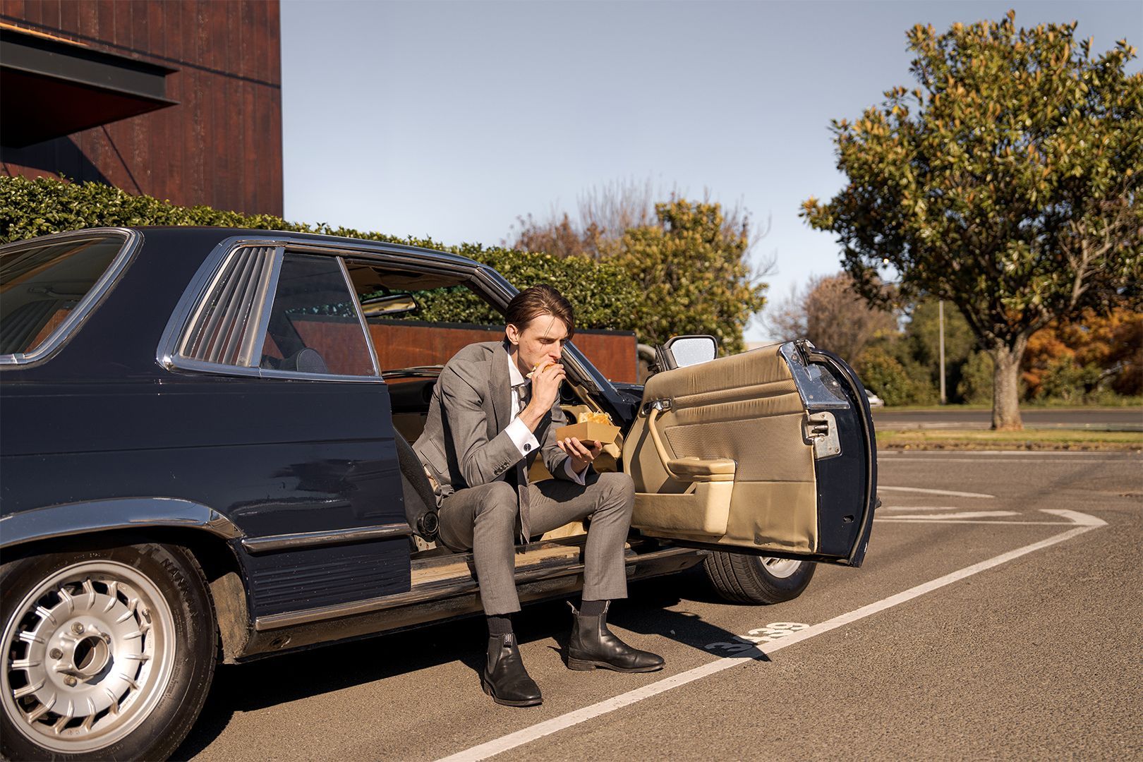 Man eating a sandwich, sitting in a vintage car with the door open 