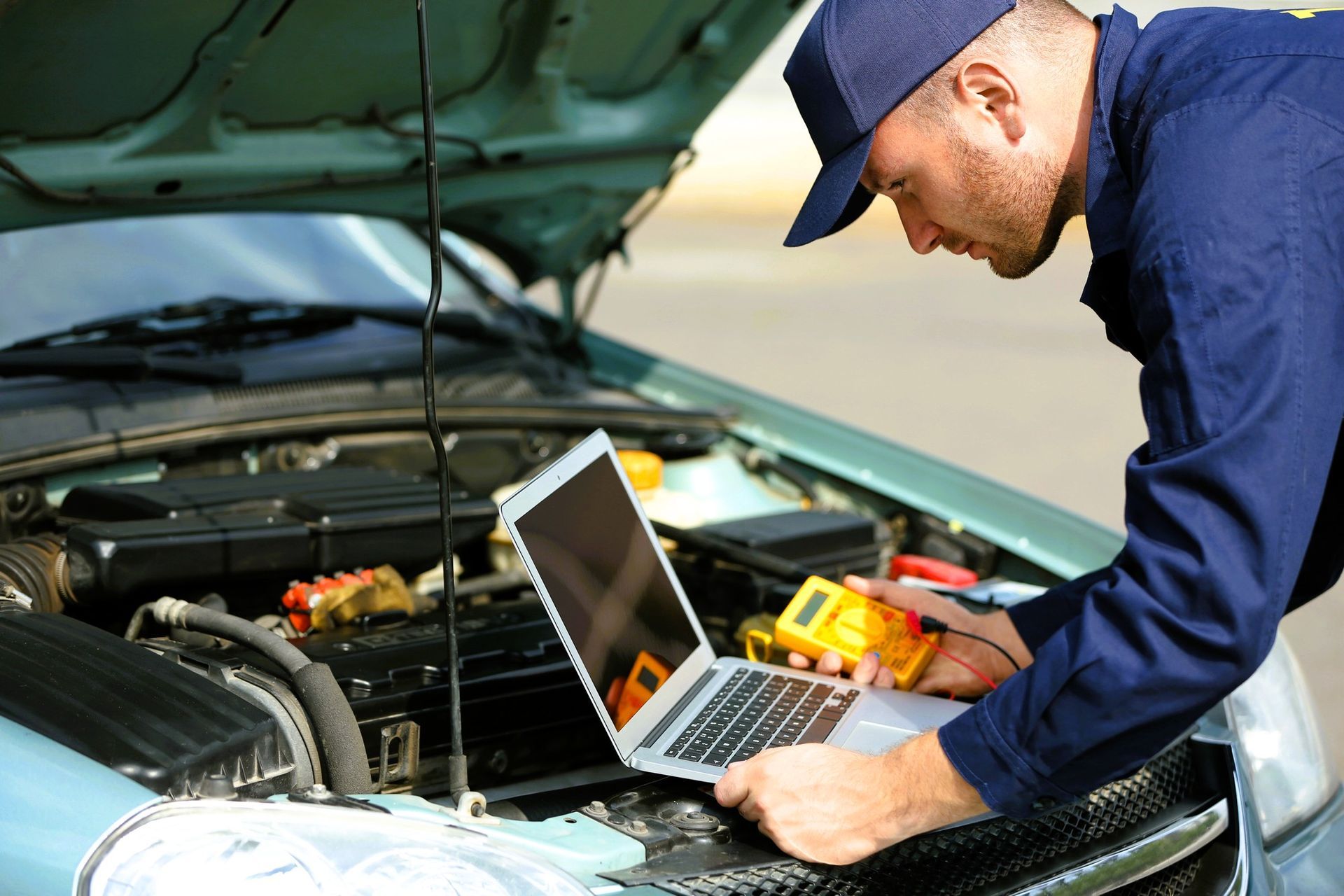 Mechanic using a laptop and multimeter to diagnose a vehicle engine under the hood
