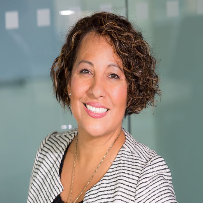 A woman with curly hair is smiling for the camera in front of a glass wall.