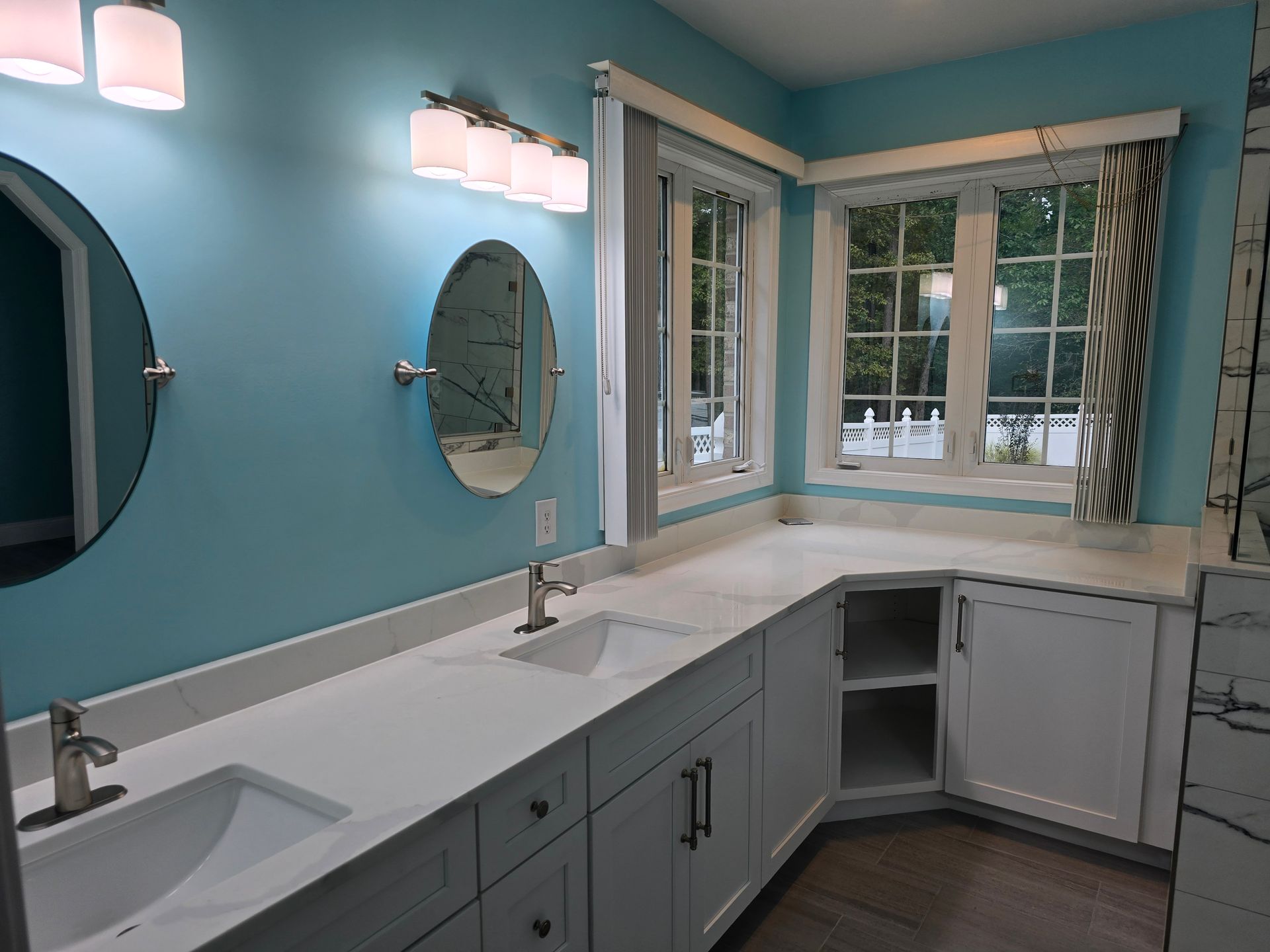 Bathroom with light blue walls, white cabinets and countertop, two sinks, and windows.