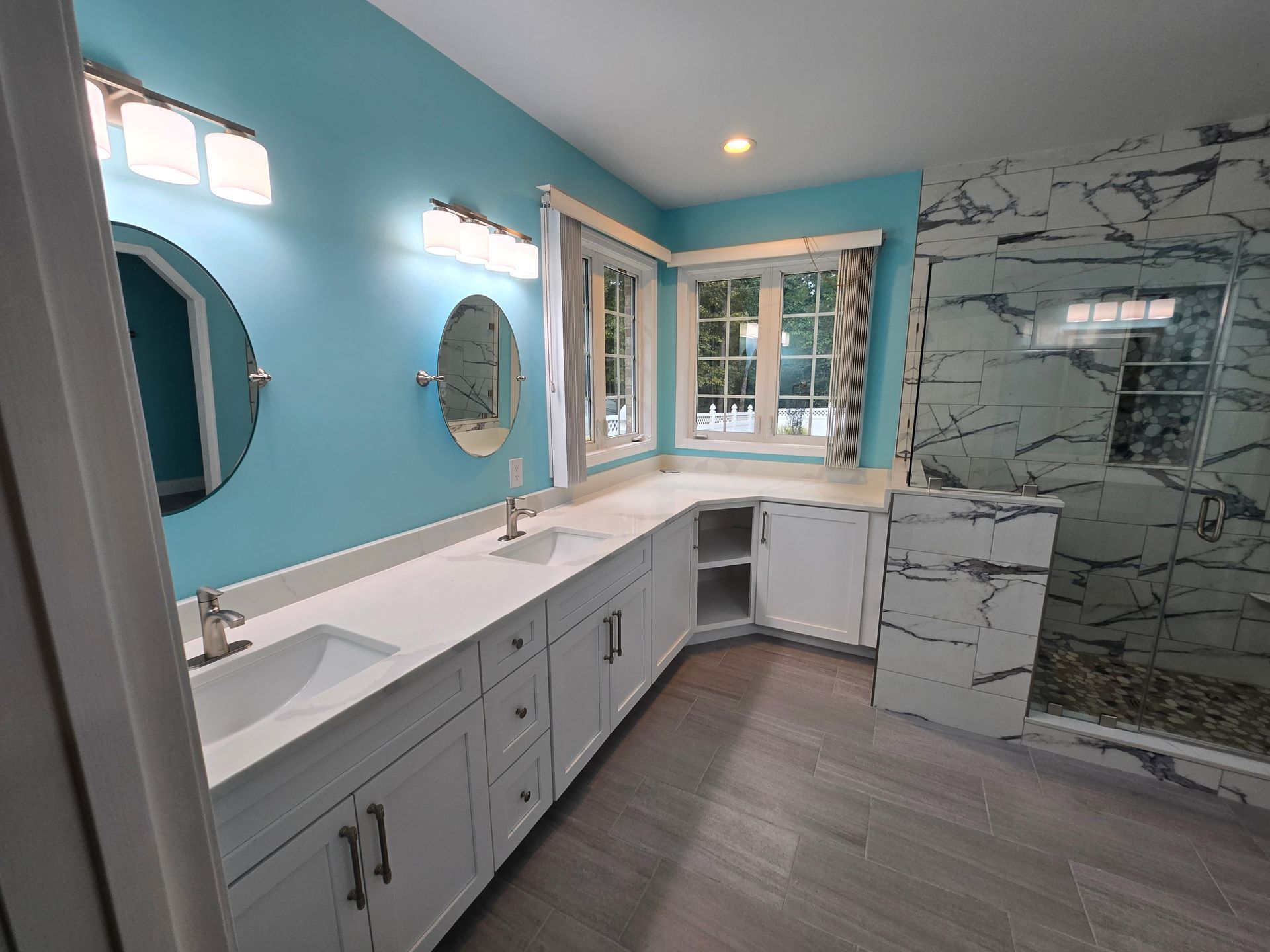Bathroom with turquoise walls, white vanity, marble shower, and two round mirrors.