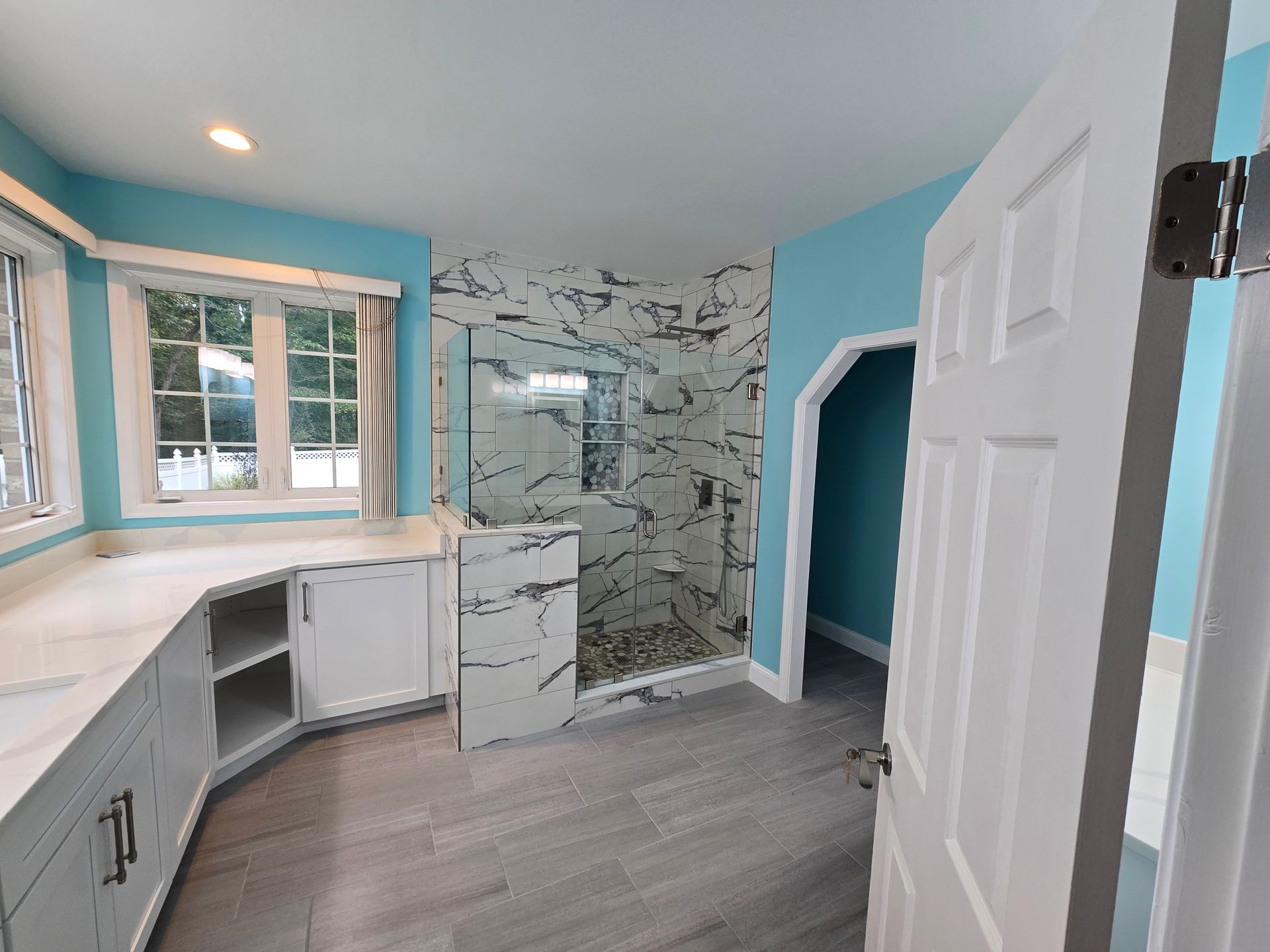 Bathroom renovation with blue walls, marble-like shower, white cabinets, and a doorway.