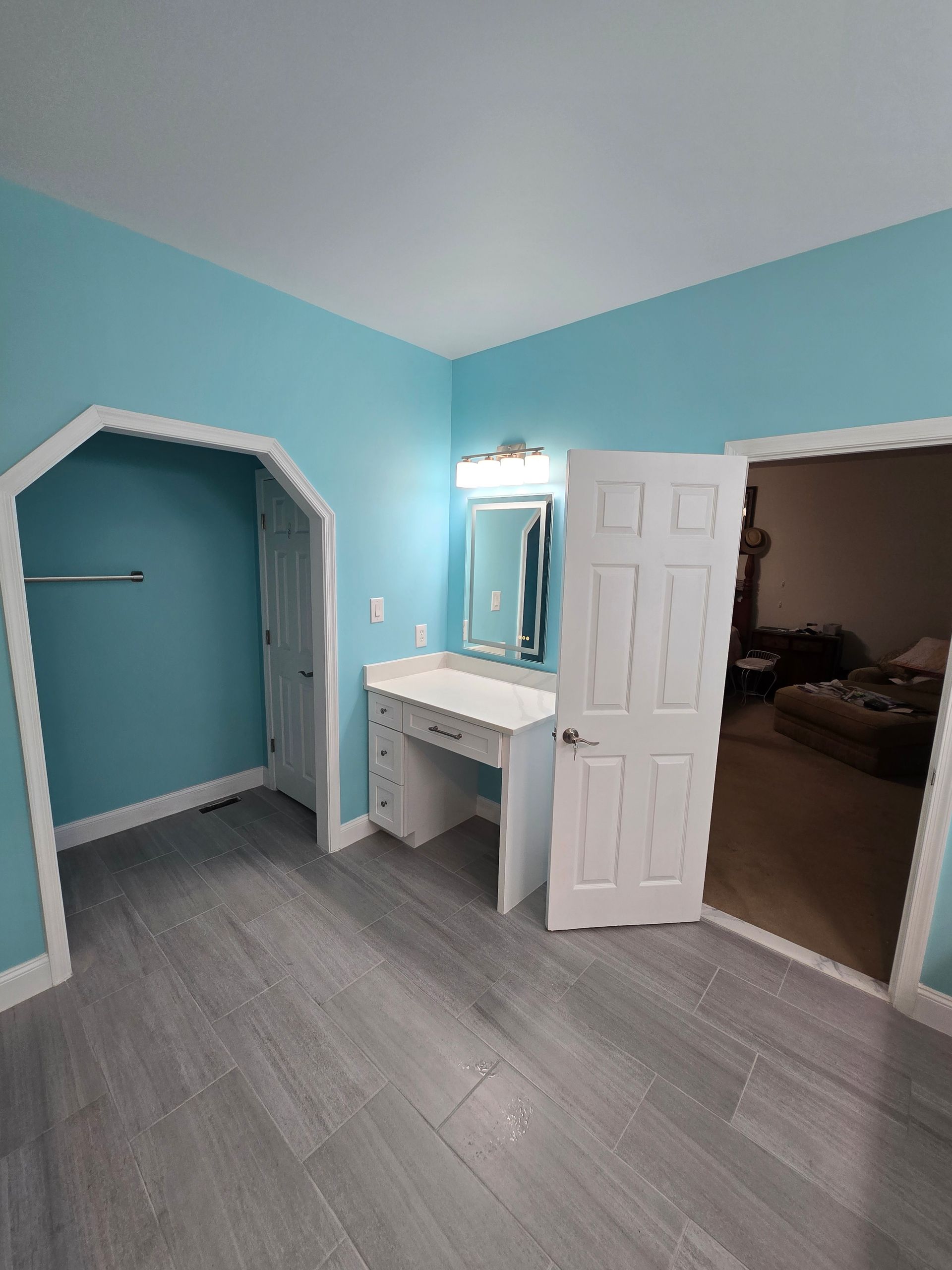 Blue-walled bedroom with white trim, dressing table, closet, and a doorway leading to another room. Gray flooring.