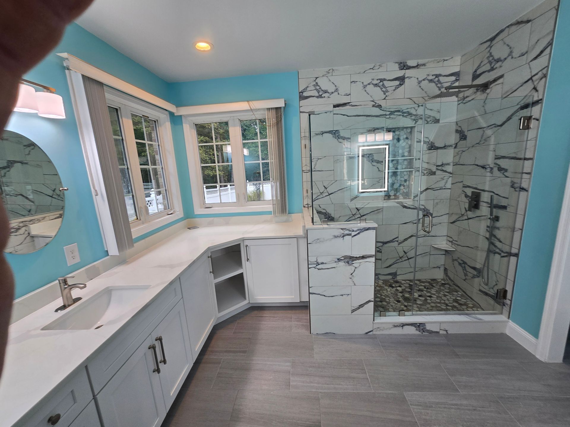 Bathroom with blue walls, white cabinets, marble shower, and a large window.