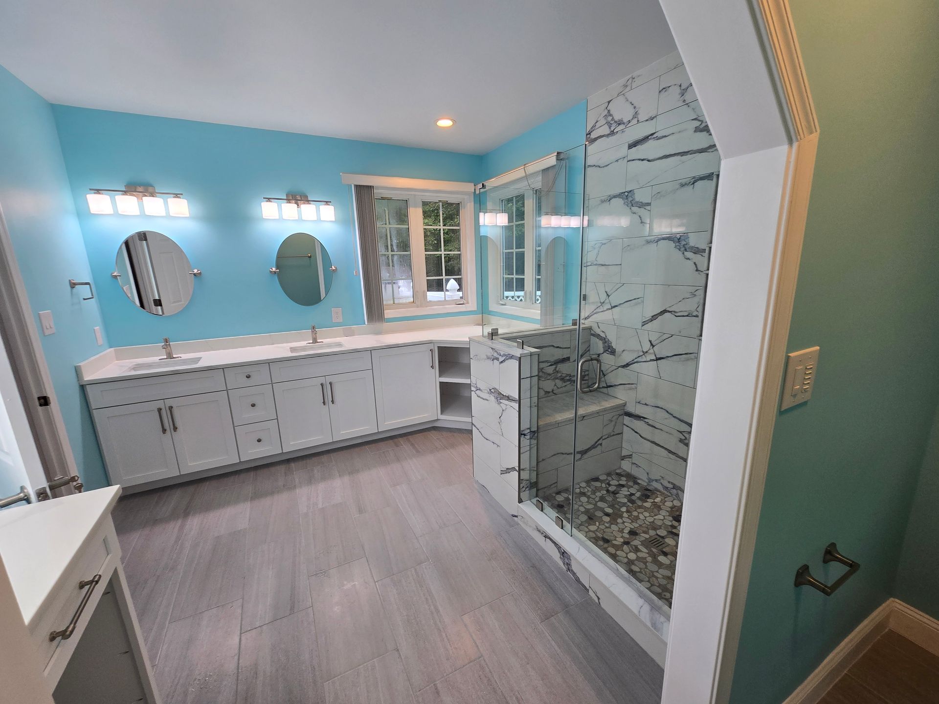 Spacious, light blue bathroom with white cabinetry, marble-tiled shower, two mirrors, and grey wood-look flooring.