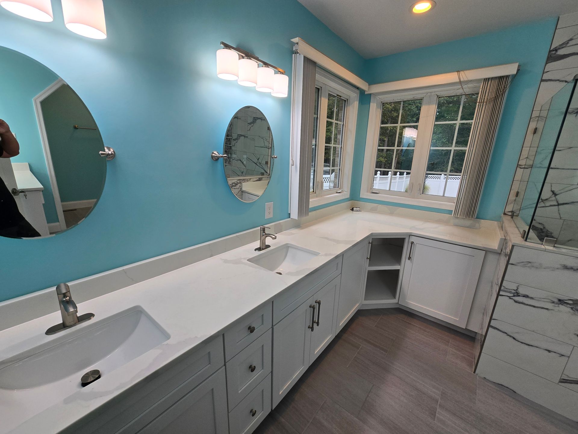 Blue bathroom with white cabinets, countertops, and oval mirrors over sinks. A window is in the corner.