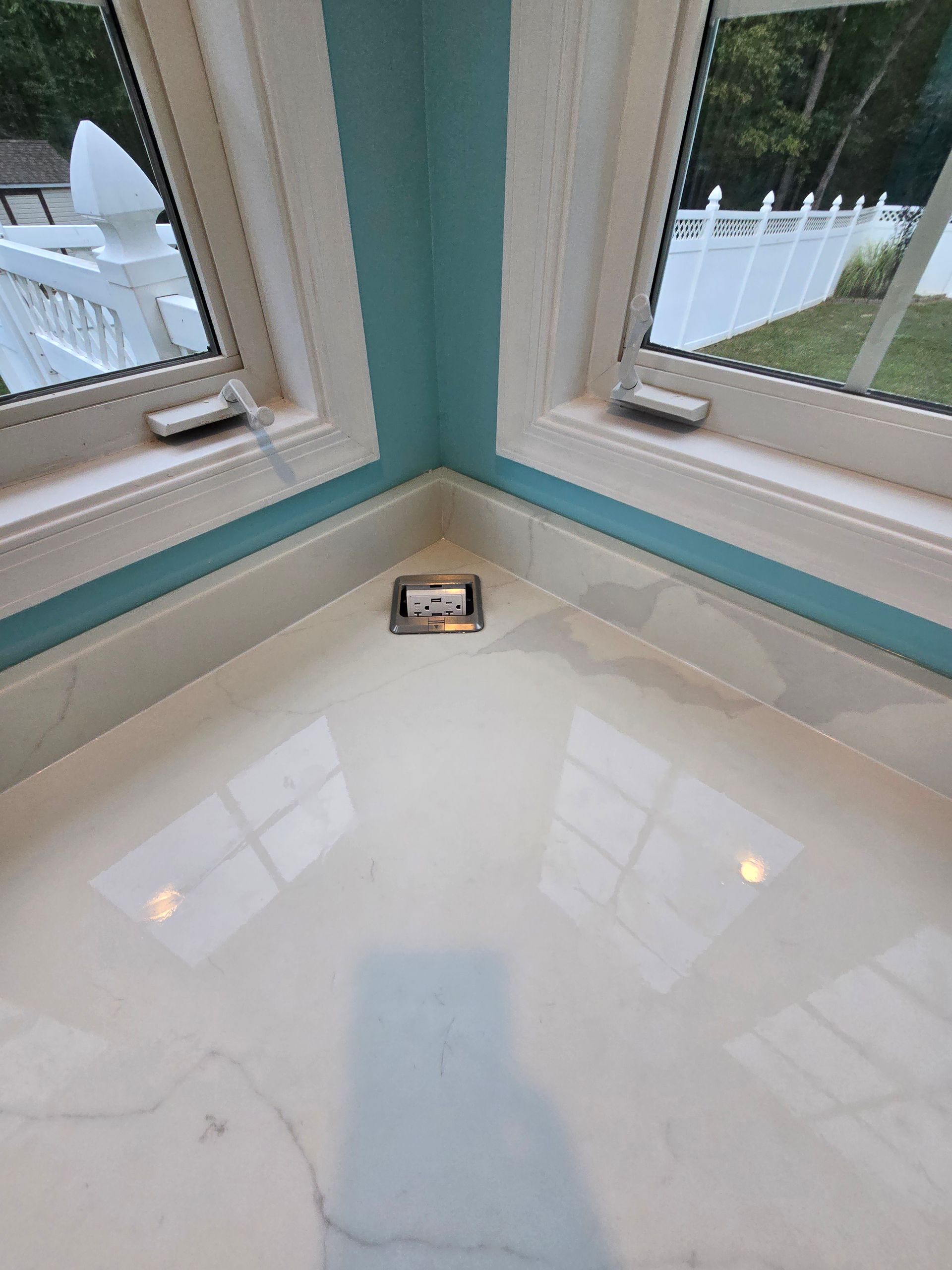 Corner countertop with light-colored surface, beneath two windows with a view of a white fence.