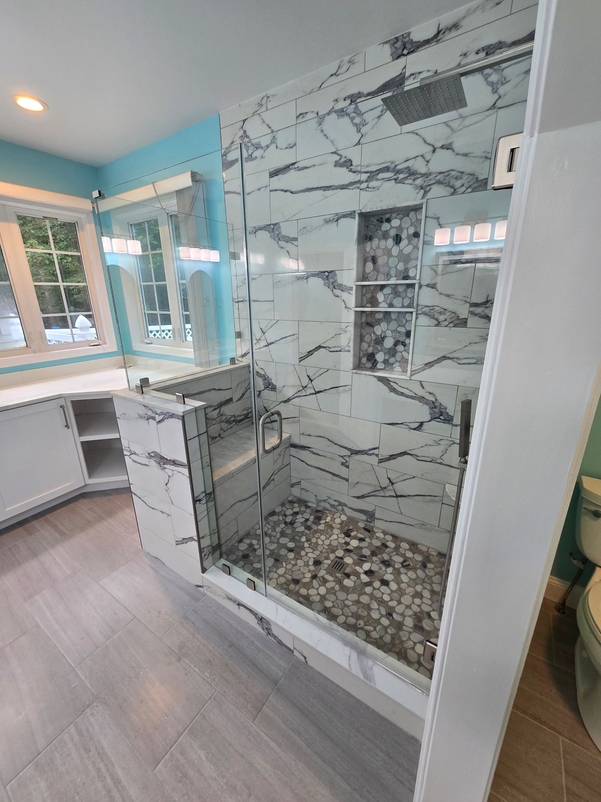 Modern bathroom with marble shower, glass door, pebble floor, white cabinets, and blue walls.
