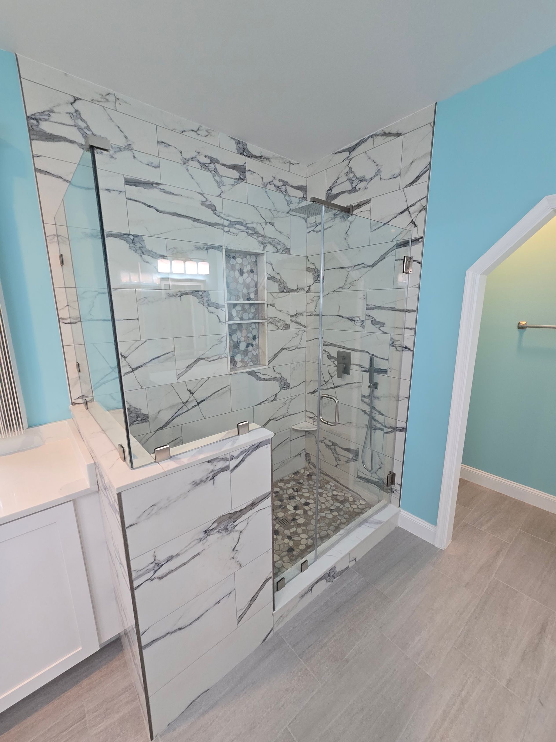Modern bathroom with marble shower, pebble floor, glass door, and turquoise walls.