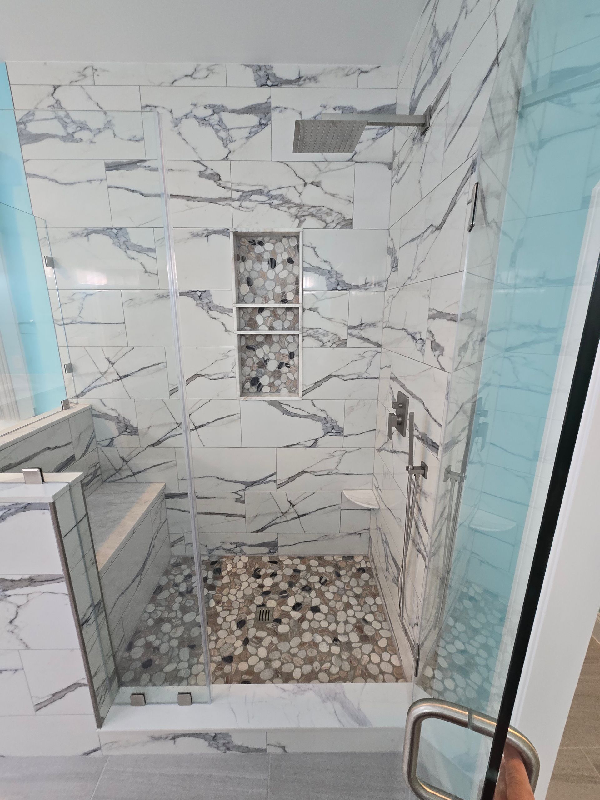 Modern shower with marble-look tile, pebble floor, glass door, and built-in niche.