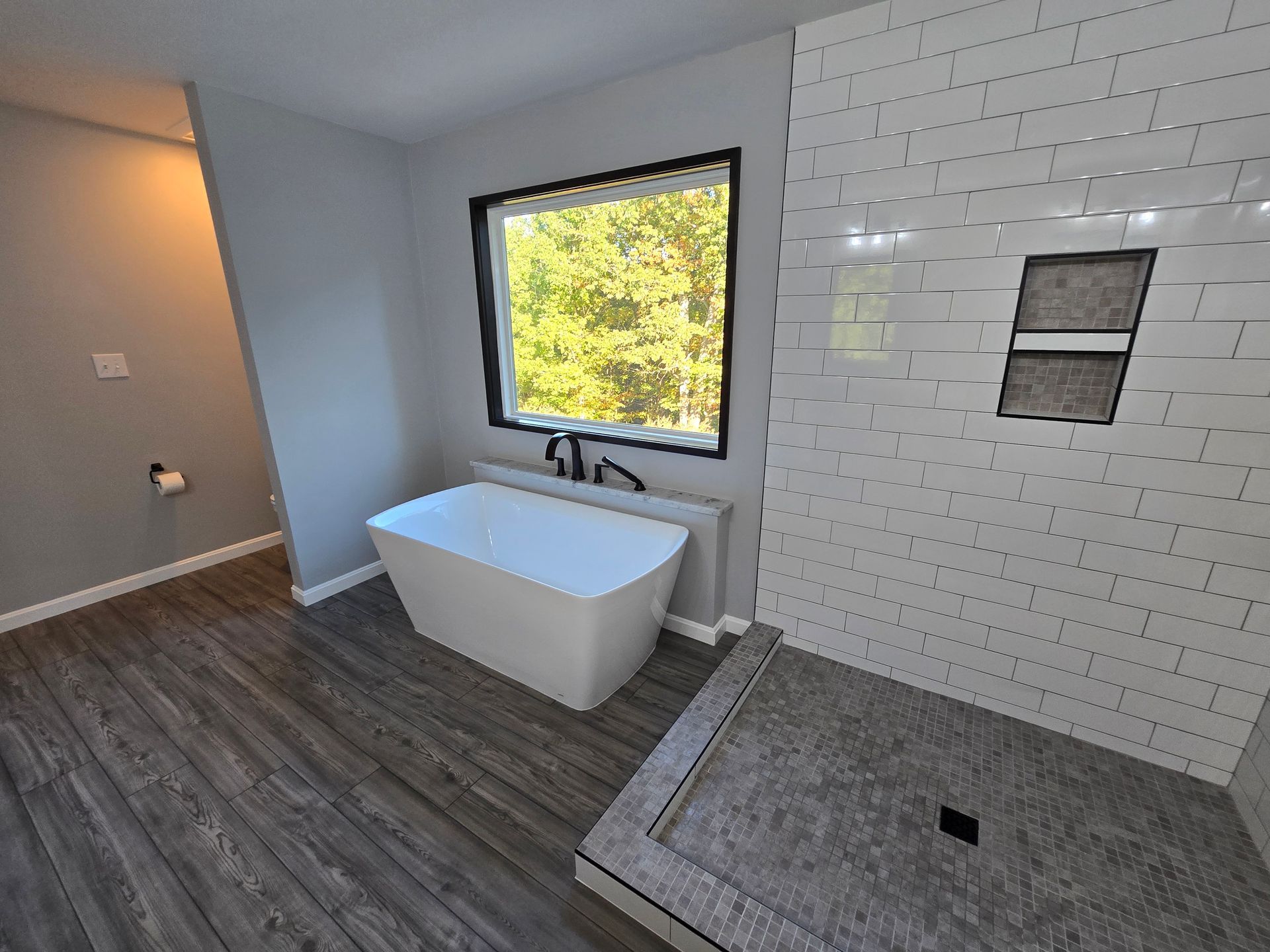 Modern bathroom with a tub, shower, window, and gray flooring. White subway tile in the shower.