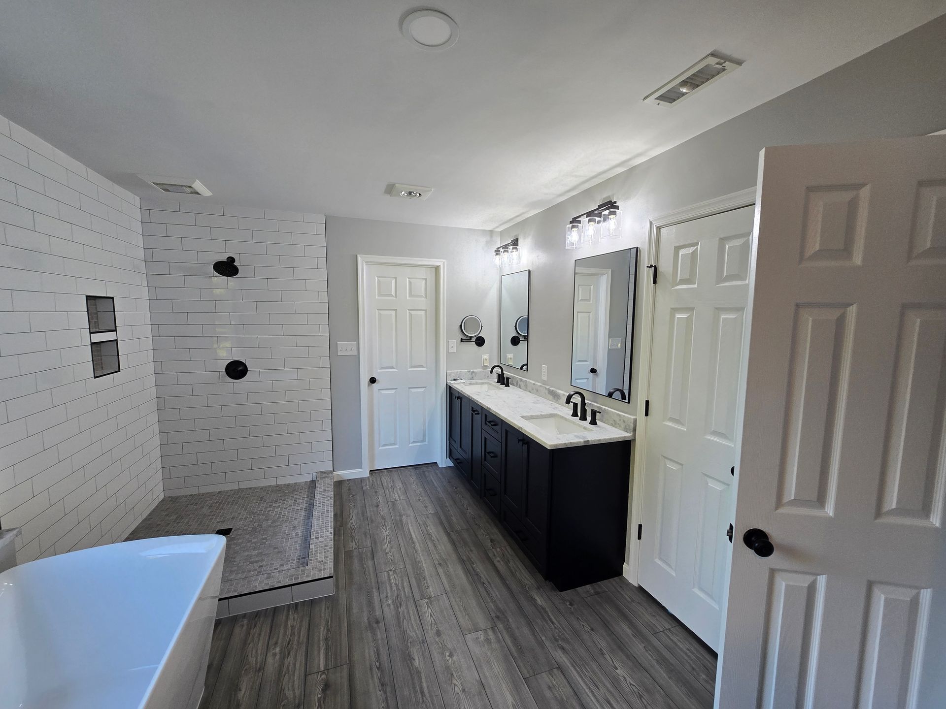 Spacious, modern bathroom with a walk-in shower, black vanity, white walls, and wood-look flooring.