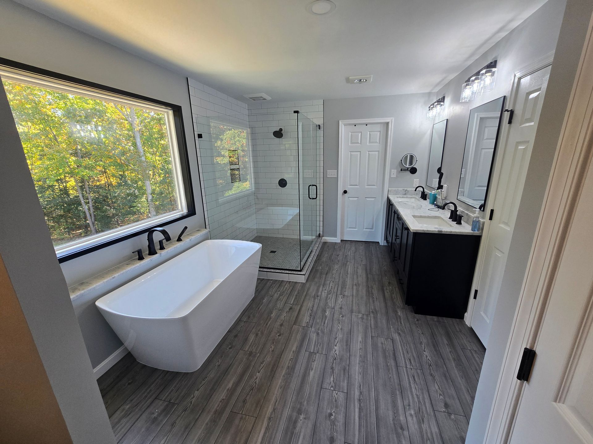 Modern bathroom with white tub, shower, and vanity. Large window with outdoor view, gray flooring.