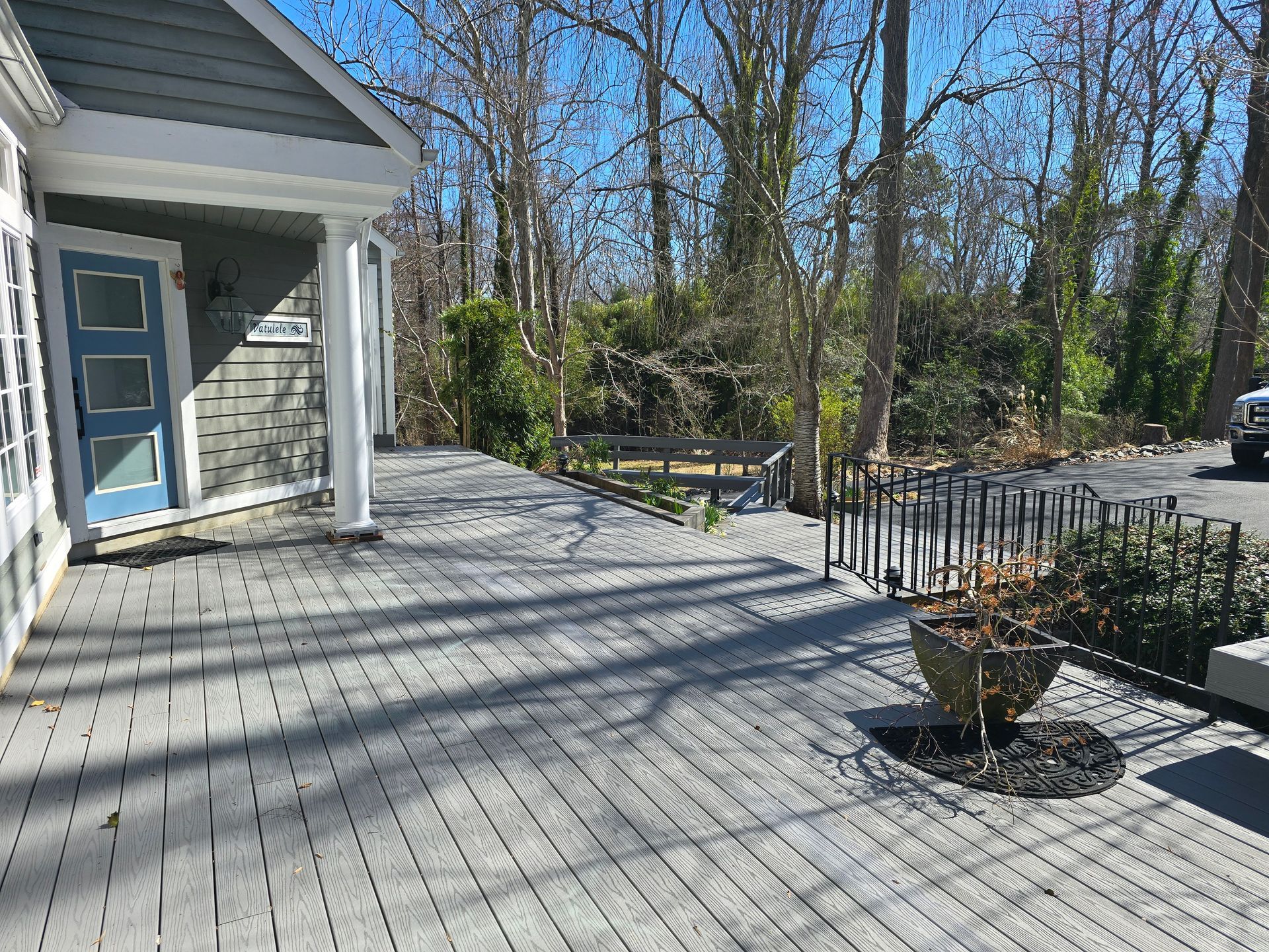 Wooden deck extends from a gray house with blue door, leading to a path with a bench, surrounded by trees.