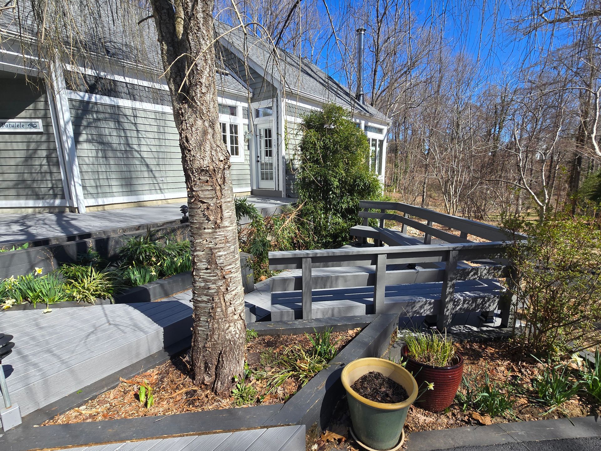 Gray house with ramp and stairs. Wooden ramp leads to the entrance. Landscaping with plants and a tree in front.