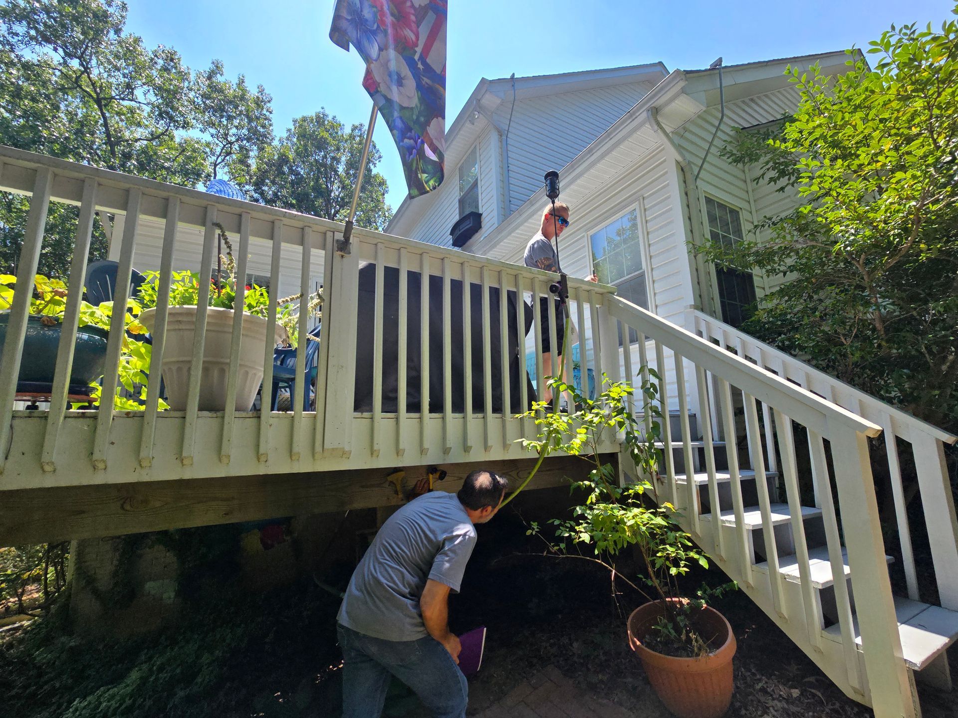 Two people working on a deck, one above the other below. White house with a flag and stairs in the background.