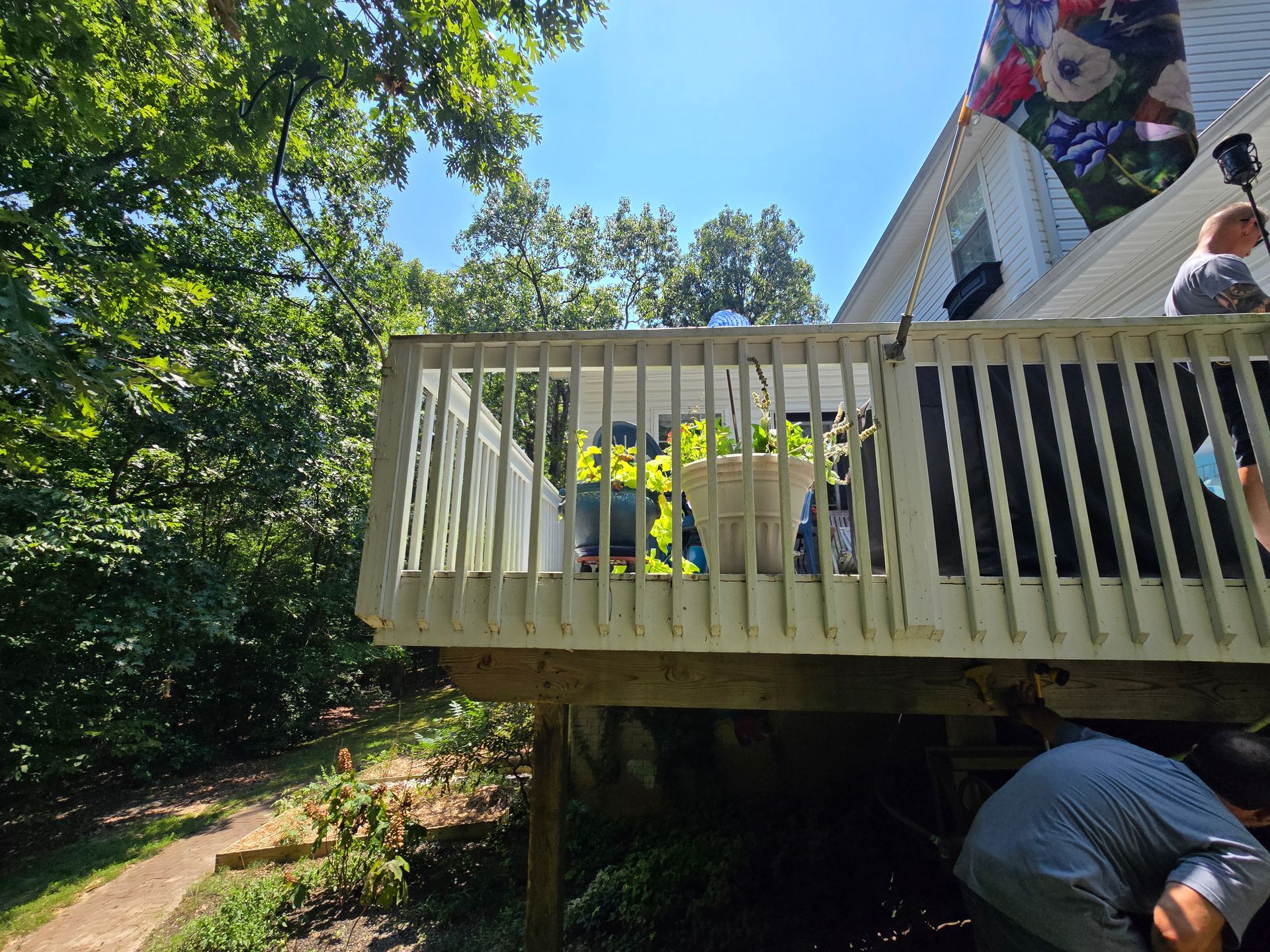 Wooden deck with potted plants, attached to a white house. Man near deck's base. Green foliage in background.