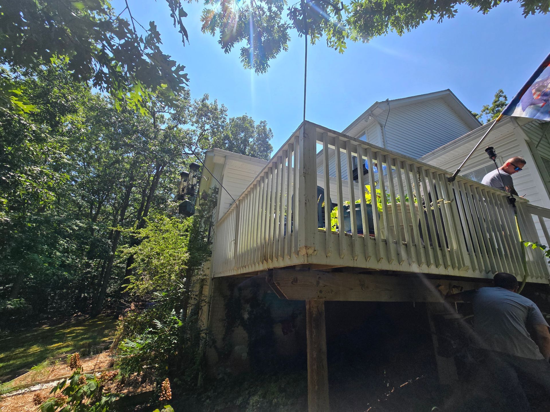 Workers power washing a wooden deck on a sunny day. Green trees surround a white house.