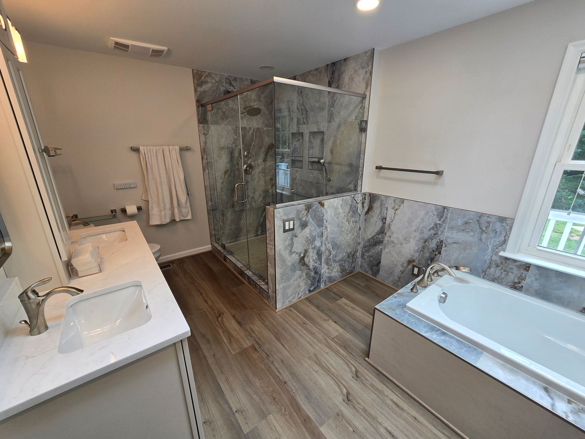 Spacious bathroom with stone-tiled shower, white vanity, and bathtub, featuring wood-look flooring and a window.