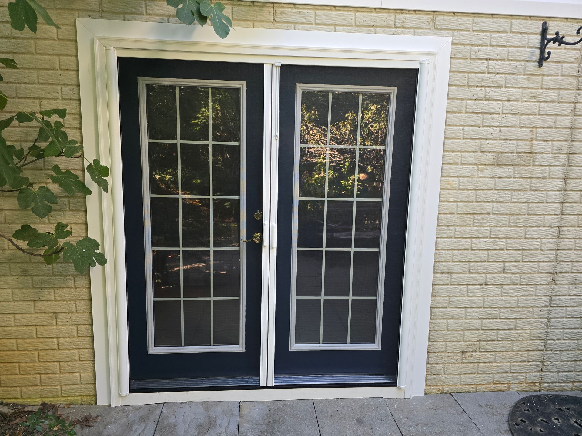 Black double doors with white grid windows and screen, surrounded by a white frame and beige brick.