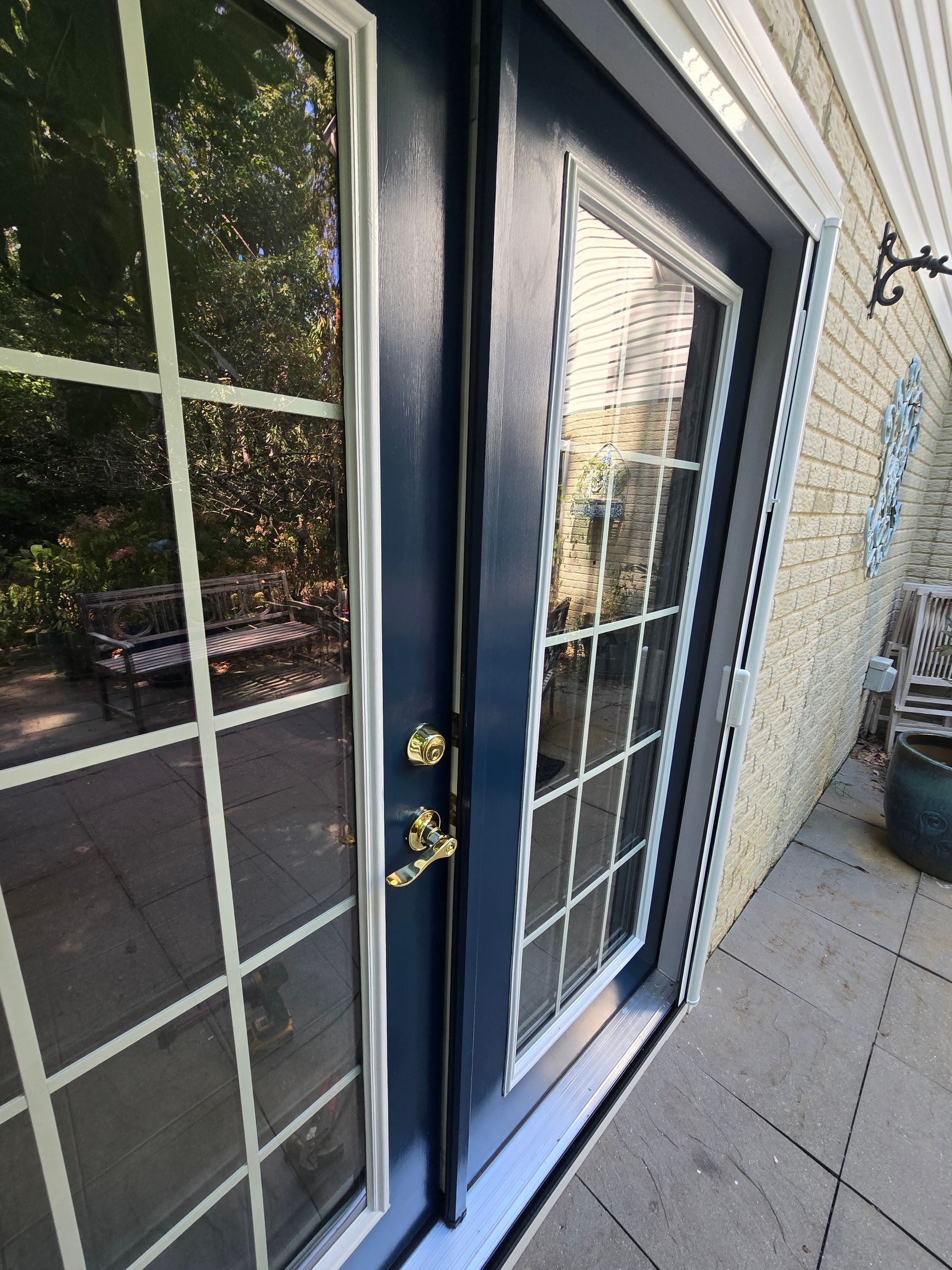 Blue French doors with gold handles reflect outdoor scene, set on a stone patio beside a brick wall.